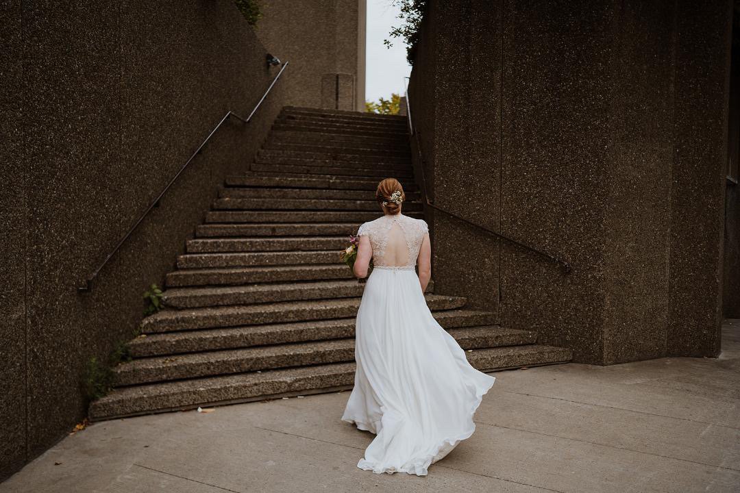 A bride in a white wedding dress walking up outdoor concrete stairs, holding a bouquet, with brown textured walls on either side and trees visible at the top of the stairs.