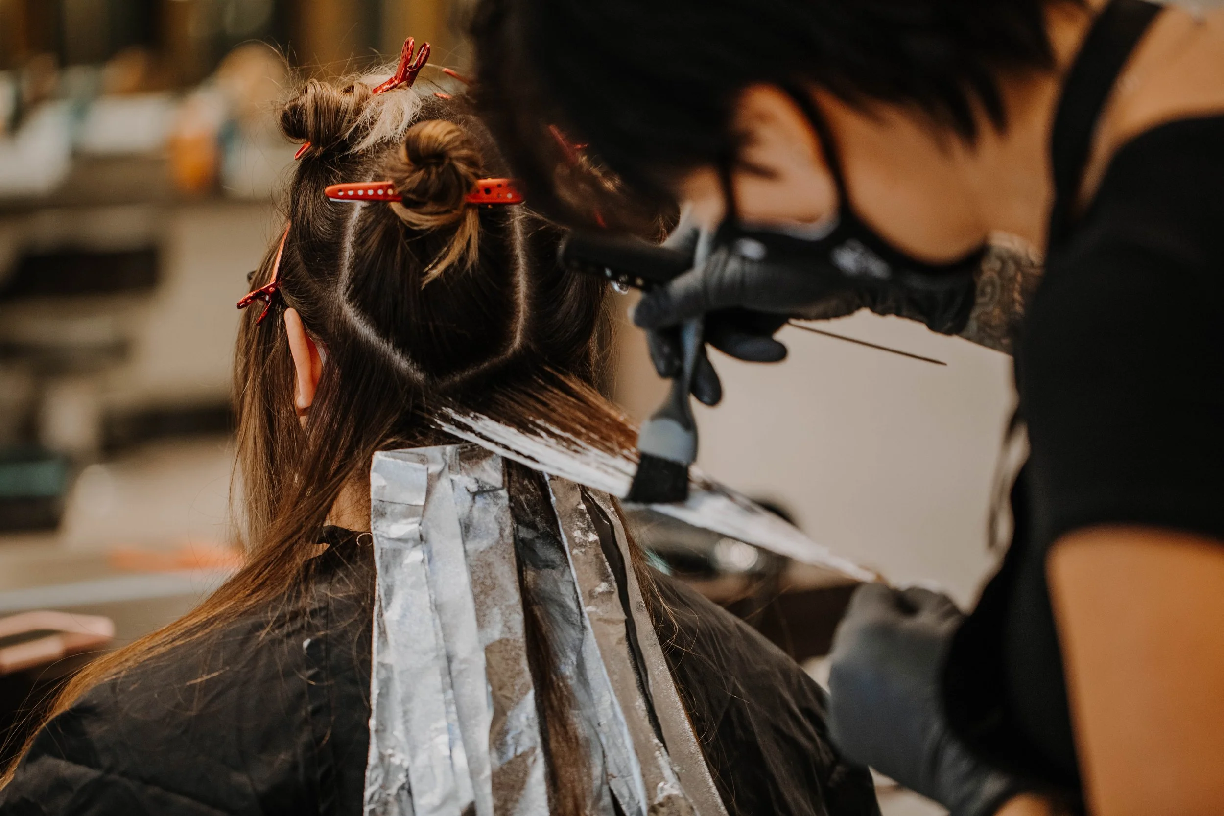 A hairstylist dyeing a woman's hair in a salon with a brush, while the woman sits with her hair sectioned and covered with foil.