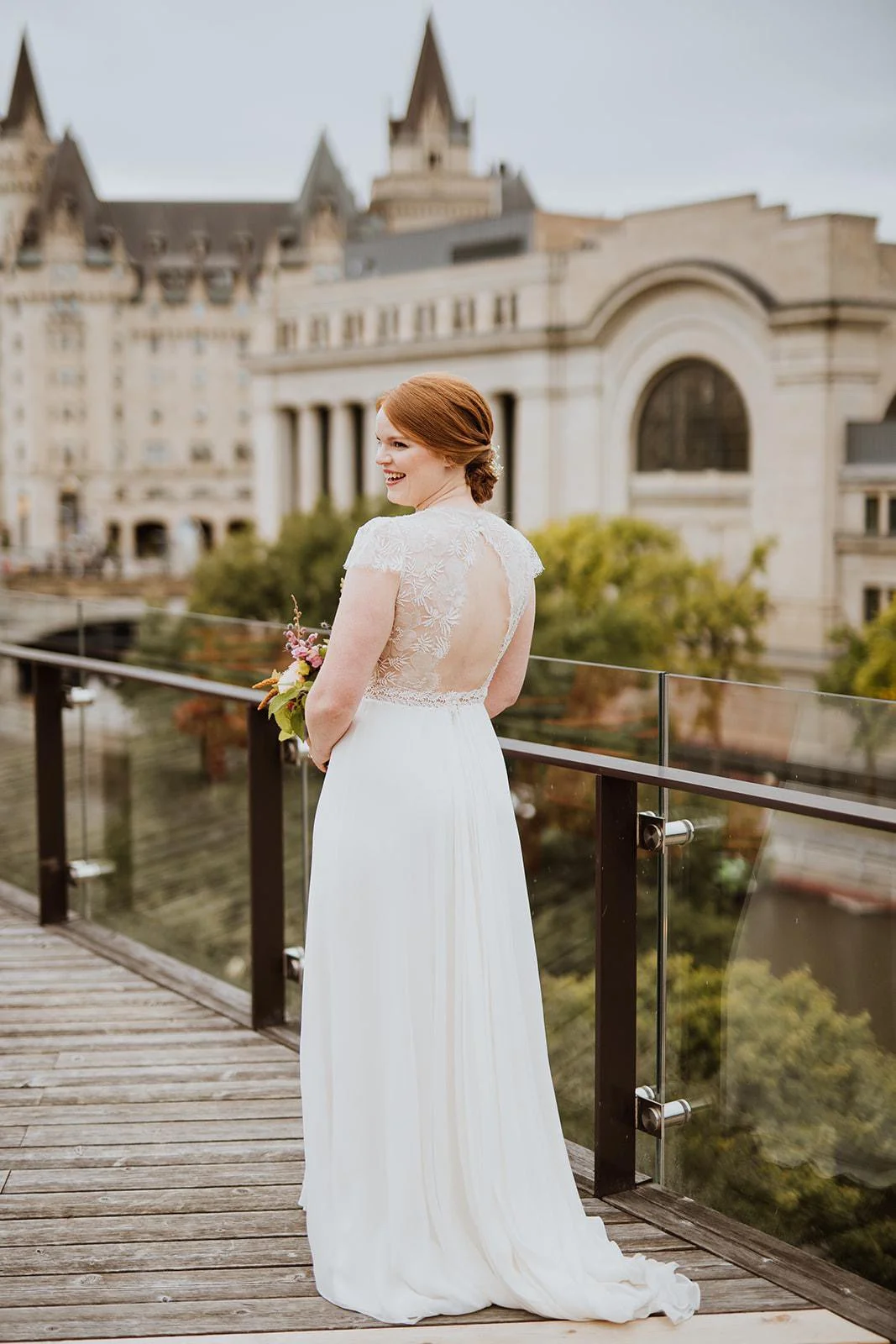 A bride with red hair in a white wedding dress with a lace back and short lace sleeves standing on a wooden balcony, holding a bouquet, and smiling, with a historic city building in the background.
