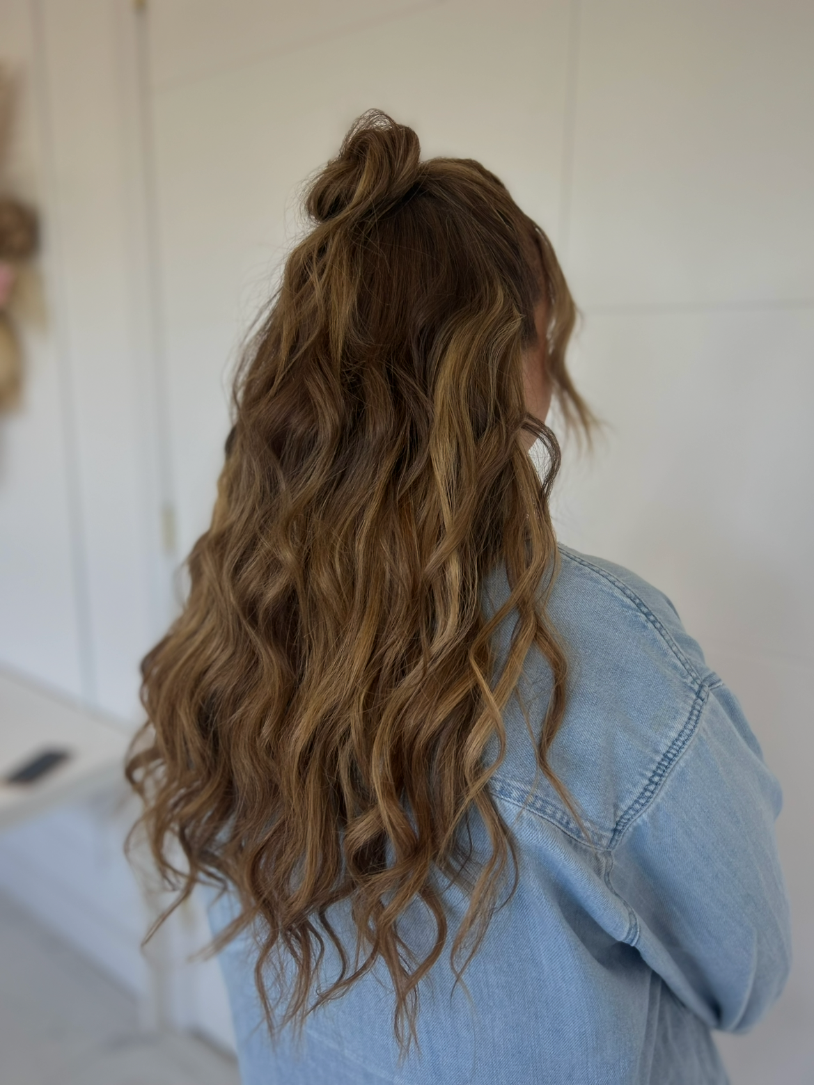 Woman with long, wavy, light brown hair wearing a denim jacket, facing away in a light-colored room.