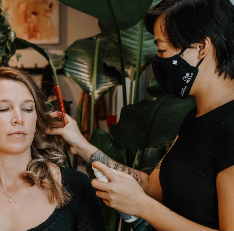 A woman with curly blonde hair gets her hair styled by a hairdresser wearing a black mask.
