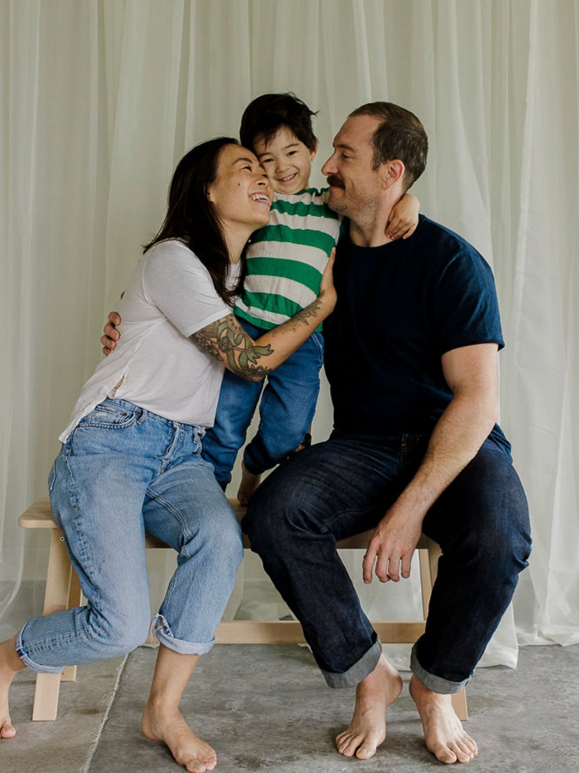 Family of three enjoying a joyful moment together, with the mother kneeling on the floor, the father sitting on a bench, and the child standing between them, all smiling and embracing each other.