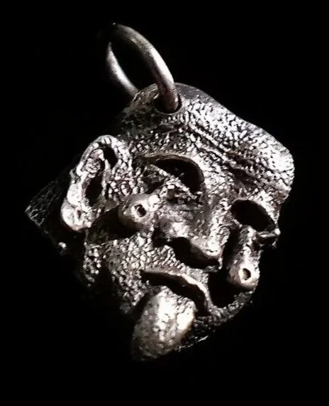 A metallic pendant resembling a human face with an open mouth and tongue sticking out, with a loop at the top for hanging.