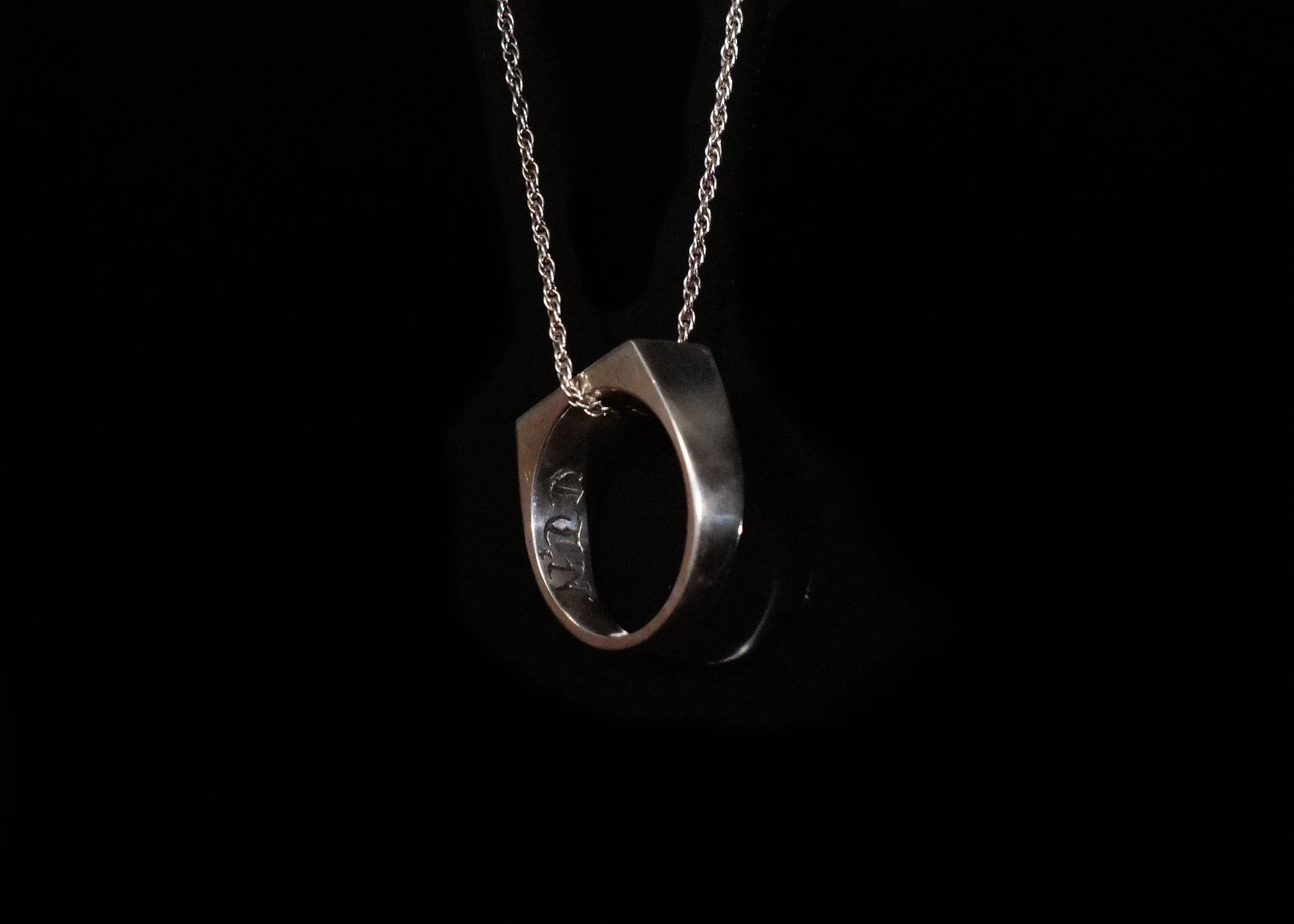 A silver ring necklace with a chain, hanging against a black background.