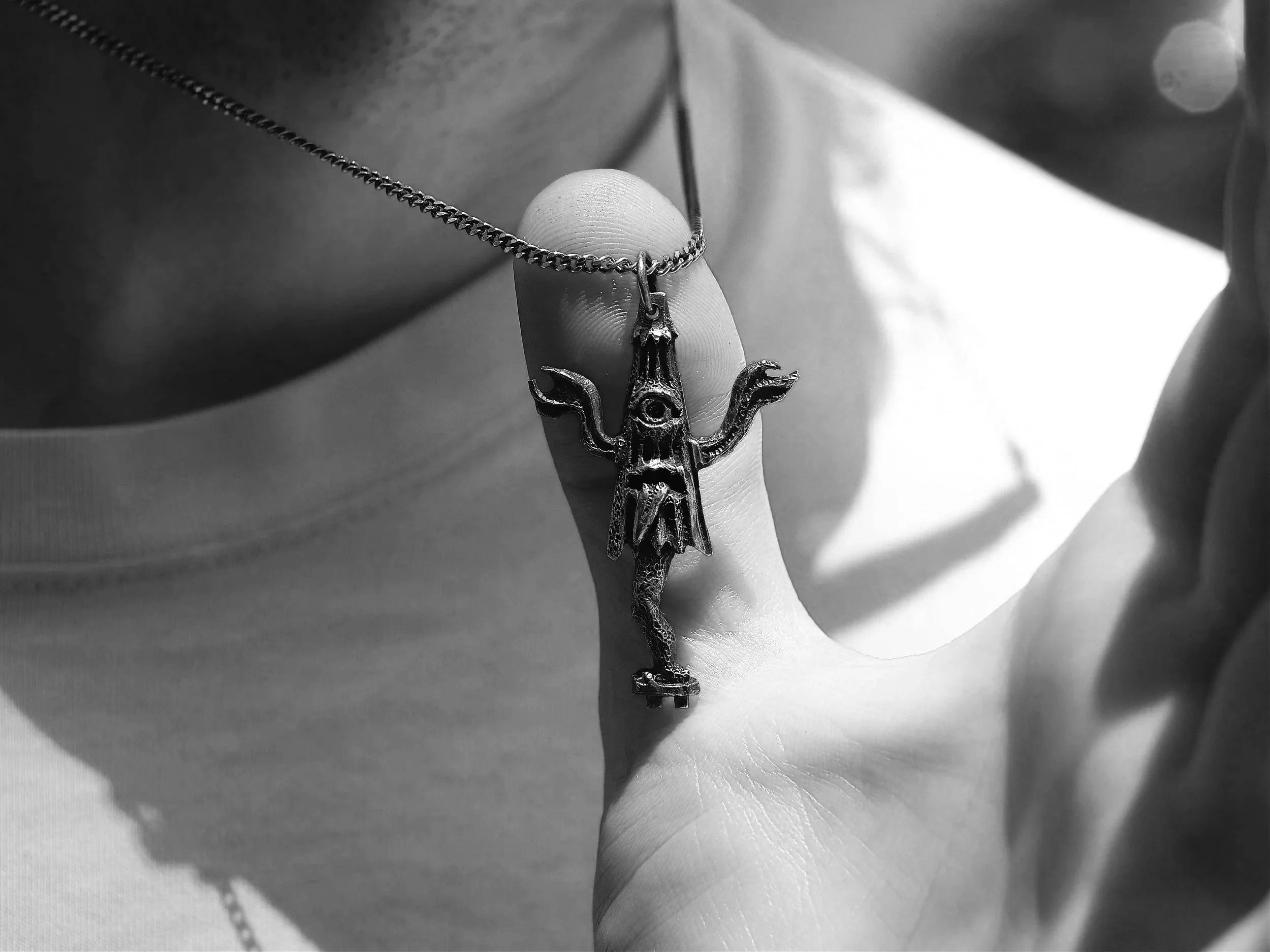 Close-up black and white photo of a hand holding a necklace with a pendant shaped like a creature with an eye and two arms.