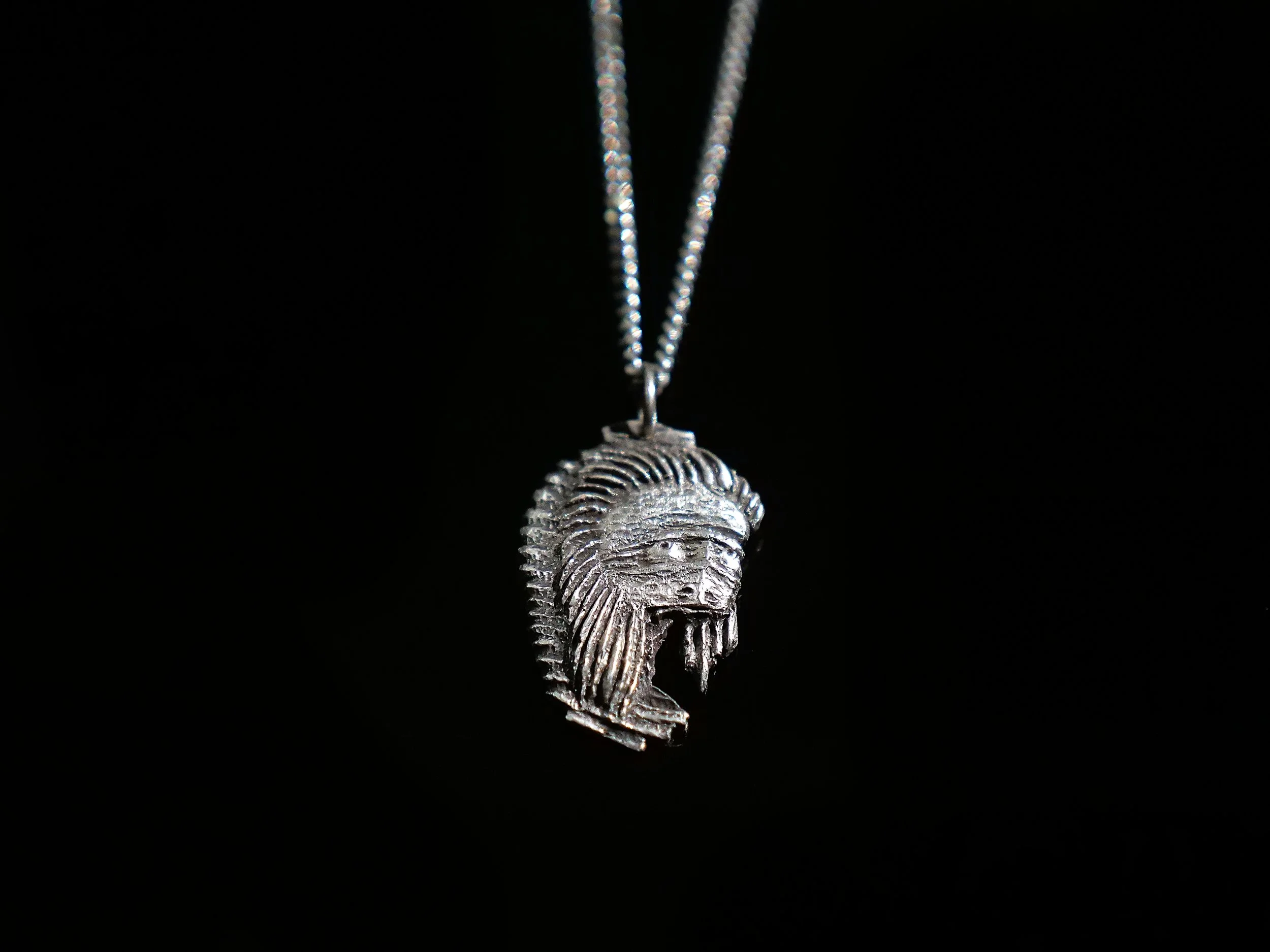 Silver necklace pendant featuring a detailed tribal headdress or warrior helmet design, hanging from a chain against a black background.