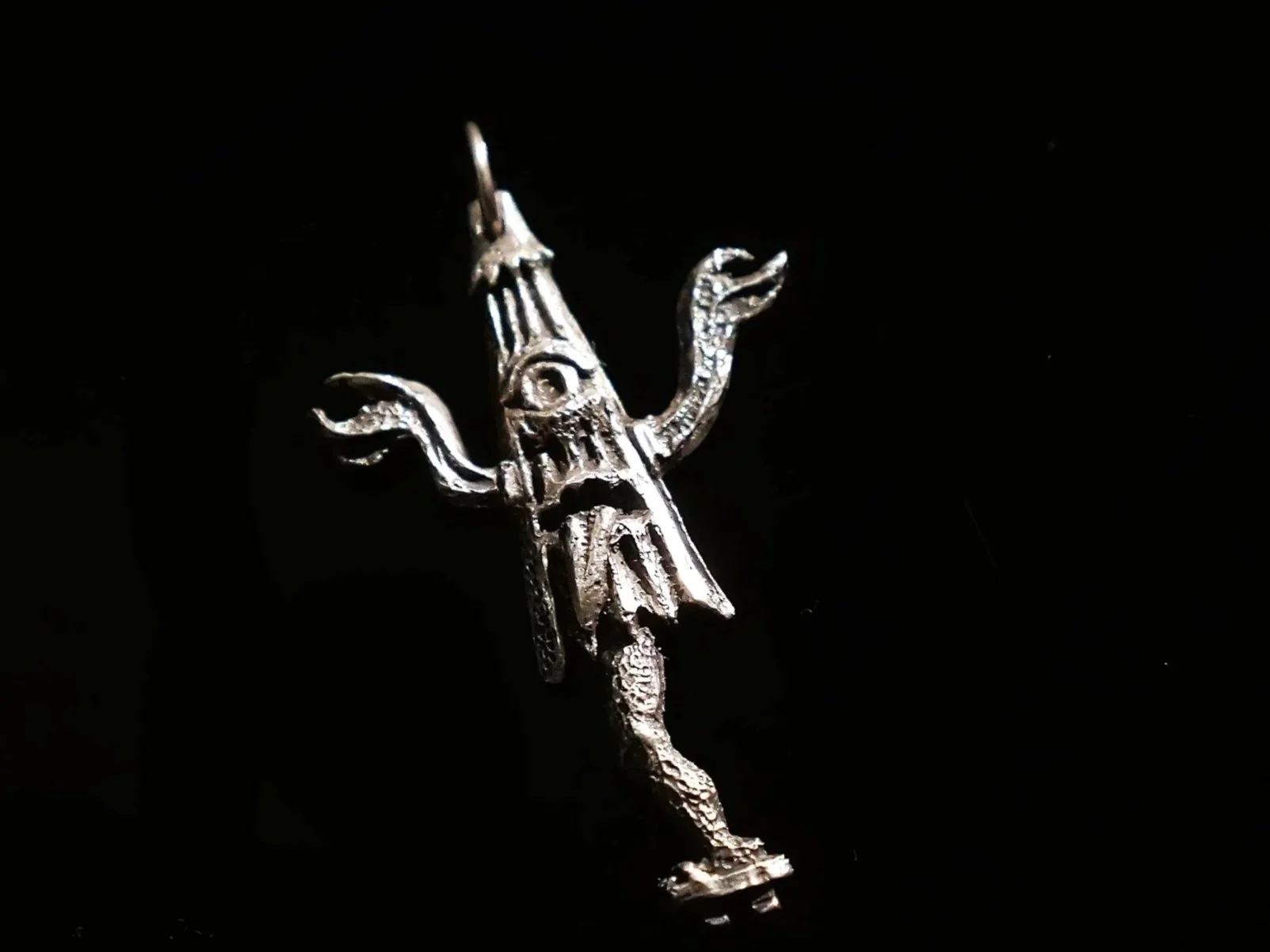 Silver pendant shaped like a mythical creature with an eye symbol, a face, and extended limbs, on a black background.