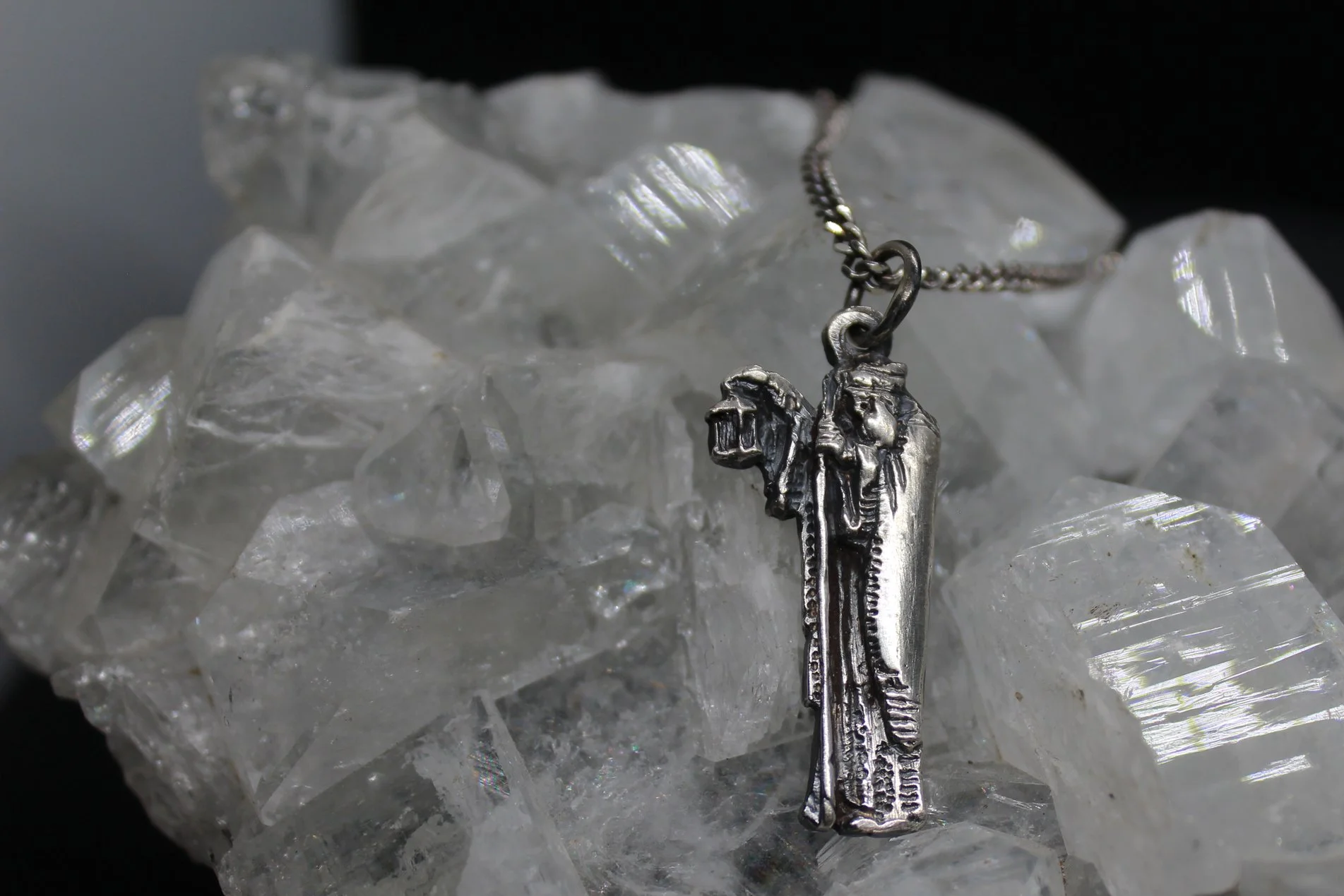 A silver necklace with a pendant featuring a detailed elongated metal figure, resting on a bed of clear, jagged ice crystals.
