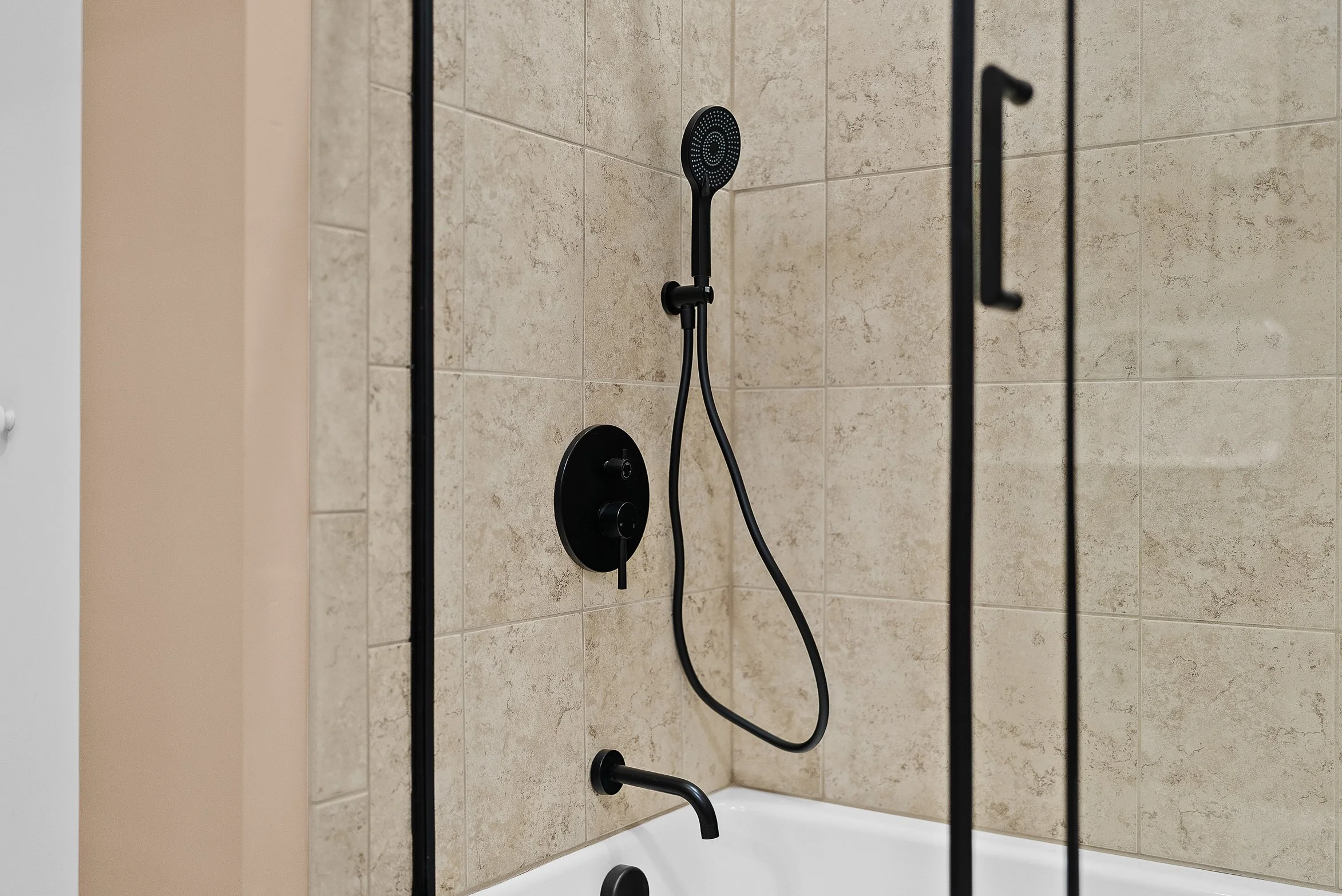 Black showerhead and fixtures in a bathroom shower with beige tiles and a glass door.