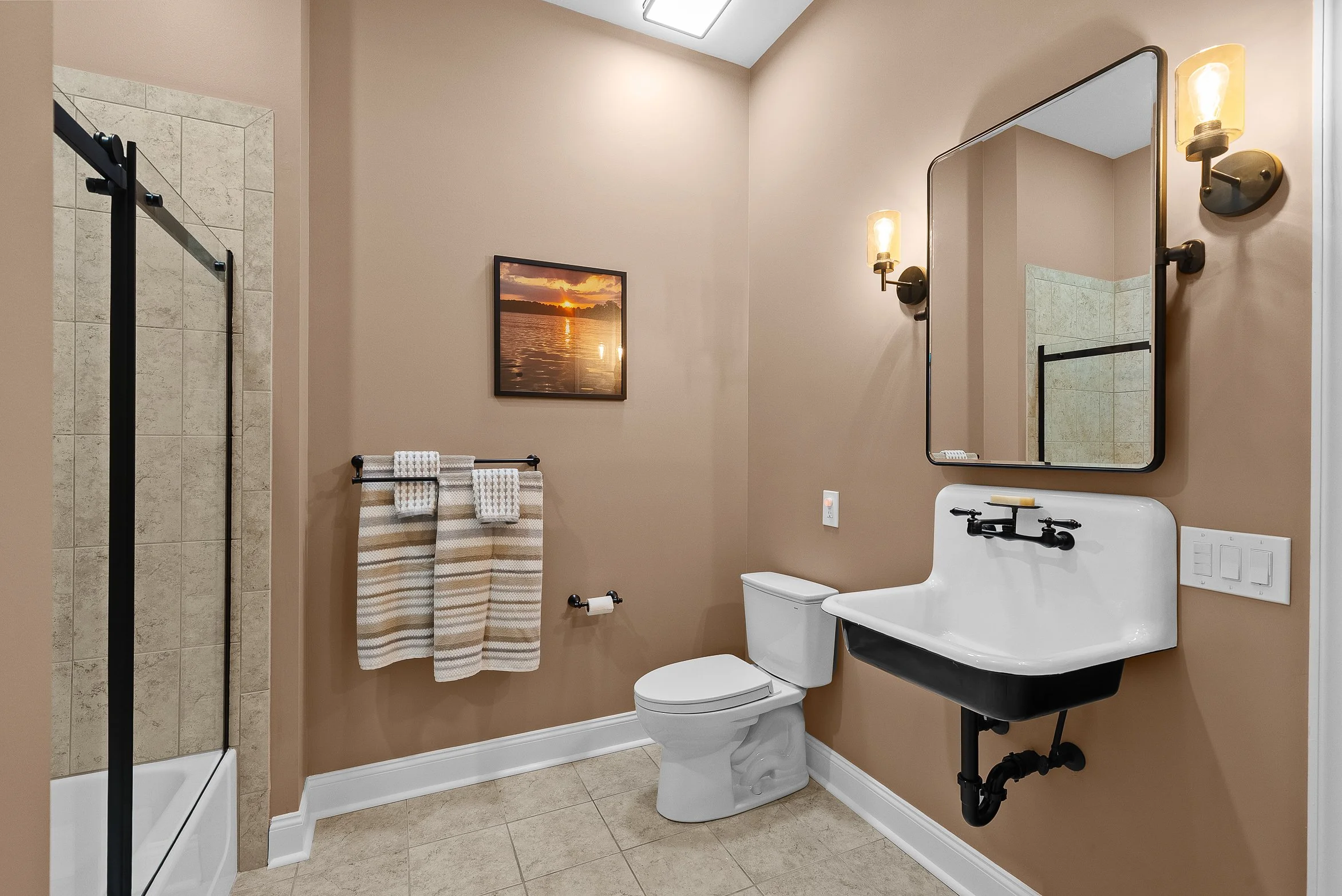 Bathroom with beige walls, tiled shower with black frame, white toilet, black sink with wall-mounted faucet, rectangular mirror, warm wall-mounted lights, framed sunset picture, striped towel on black towel rack, and beige tile floor.