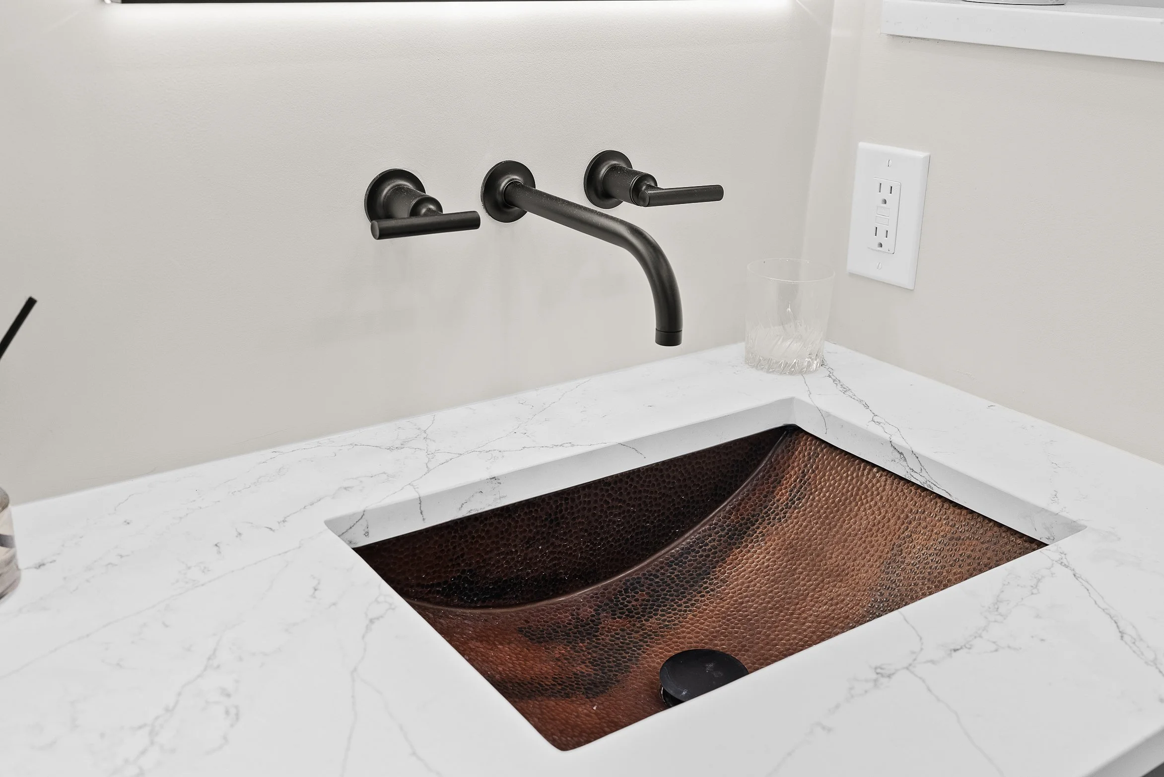 Kitchen sink with marble countertop, copper interior, black faucet, and white wall with electrical outlet
