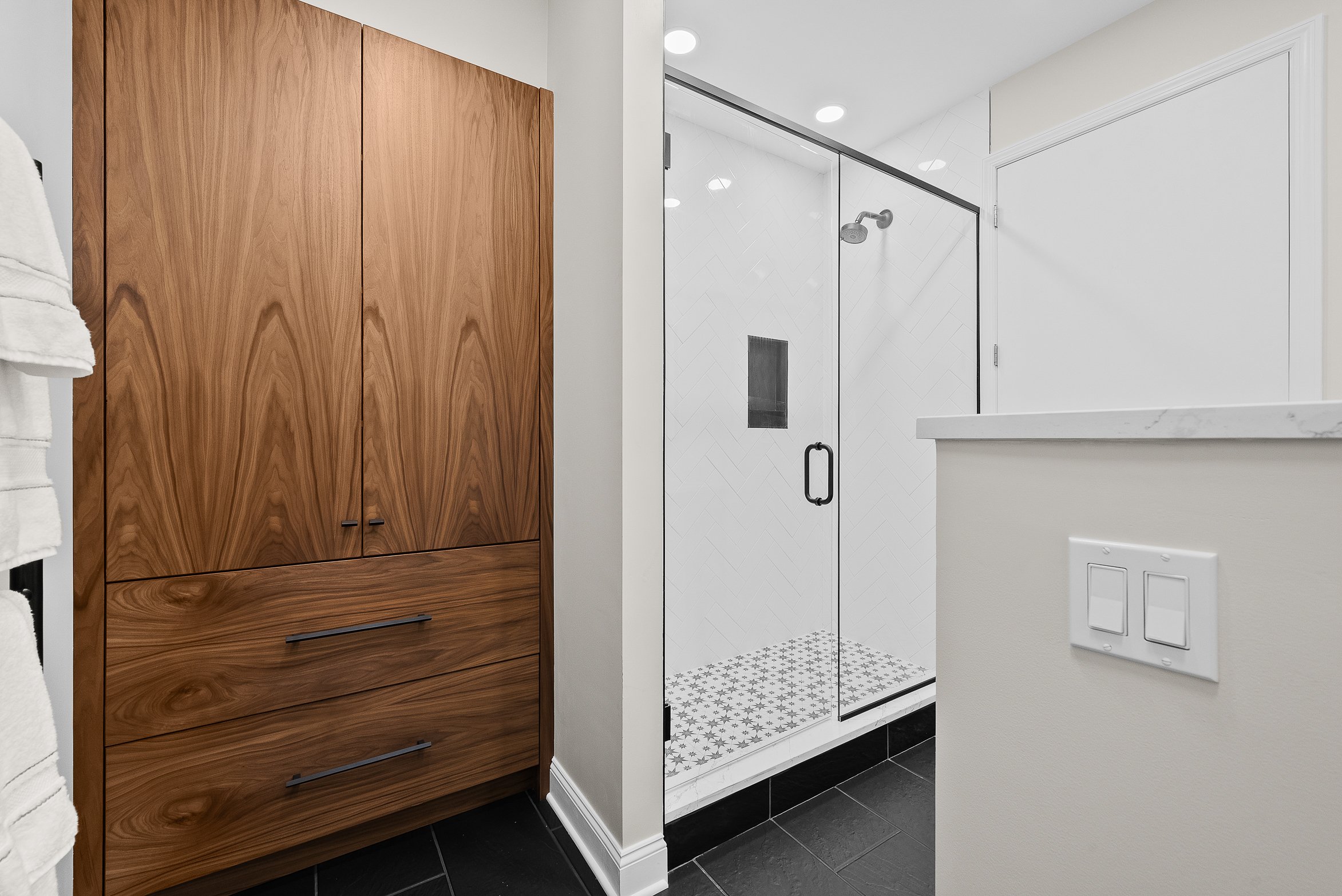 A modern bathroom corner featuring a wooden cabinet with black handles, a glass shower with white tiled walls in a herringbone pattern, a built-in shower niche, and black and white patterned floor tiles.
