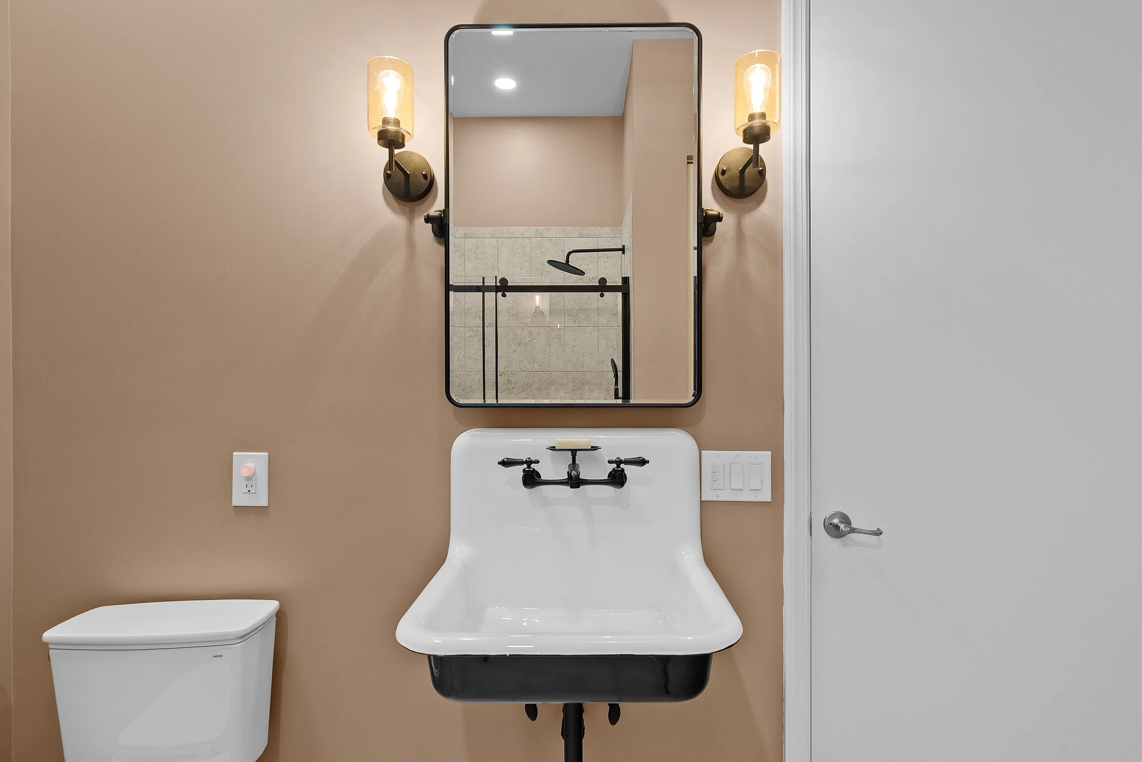 Bathroom with a beige wall, black-framed mirror, wall-mounted light fixtures, white sink with black fixtures, a white toilet, electrical outlet, and a closed white door.