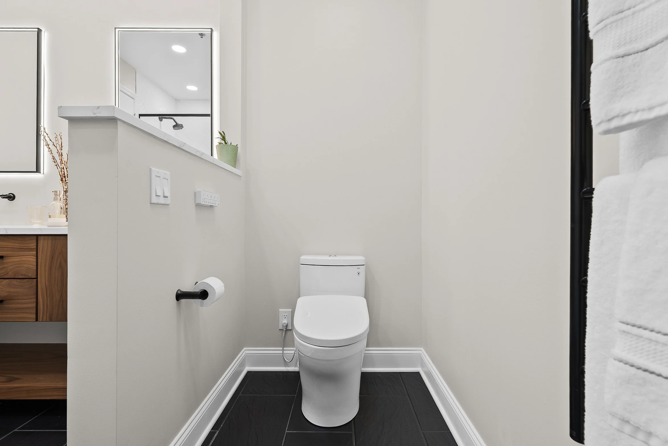 A modern bathroom with a white toilet, black tiled floor, and a partial view of a wooden vanity with a white sink, mirror, and a small potted plant.