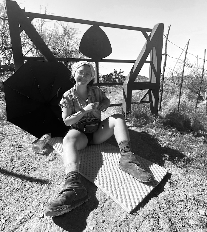 A smiling woman sitting on a textured pad on the ground outdoors, with an umbrella to her side, in front of a wooden structure with a bell and trees in the background, in black and white.