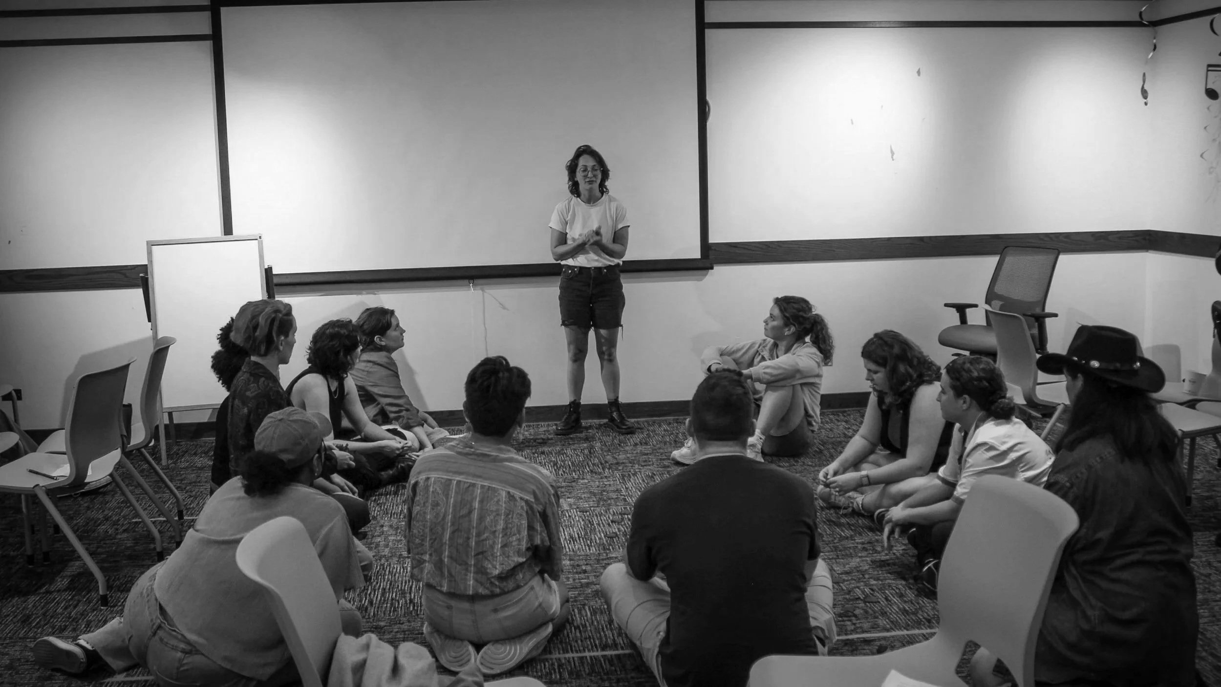 A woman standing and speaking to a group of people sitting on the floor in a conference room with a projector screen behind her.
