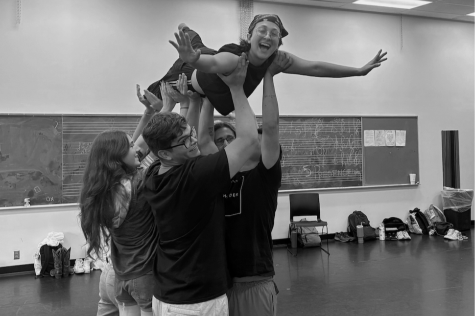 Group of people lifting a young girl in the air during a rehearsal in a classroom or dance studio with a blackboard in the background.