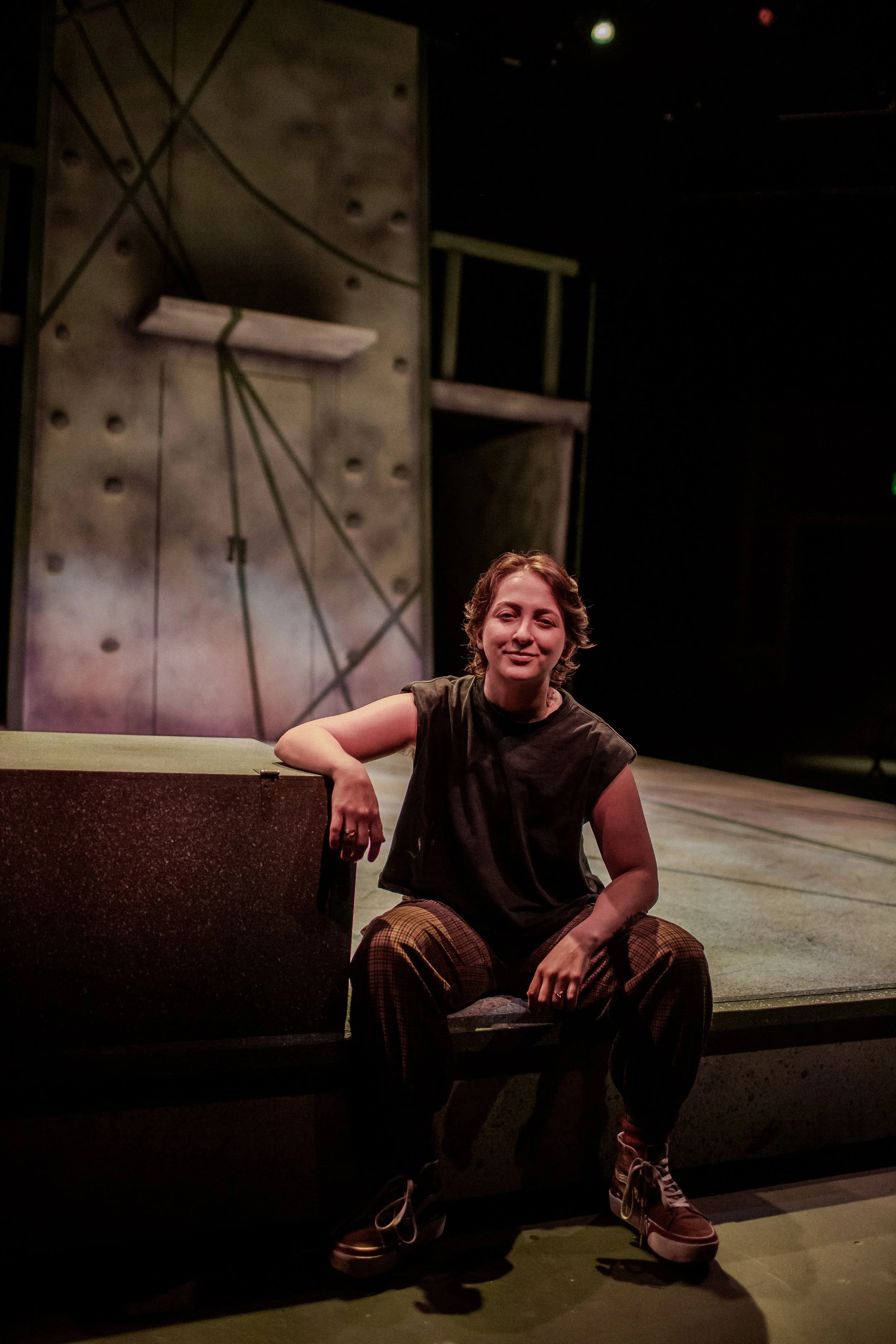 A woman with short hair and a black top sitting on a low stage with a pained expression, one arm resting on a block, in a dimly lit theater or stage setting.