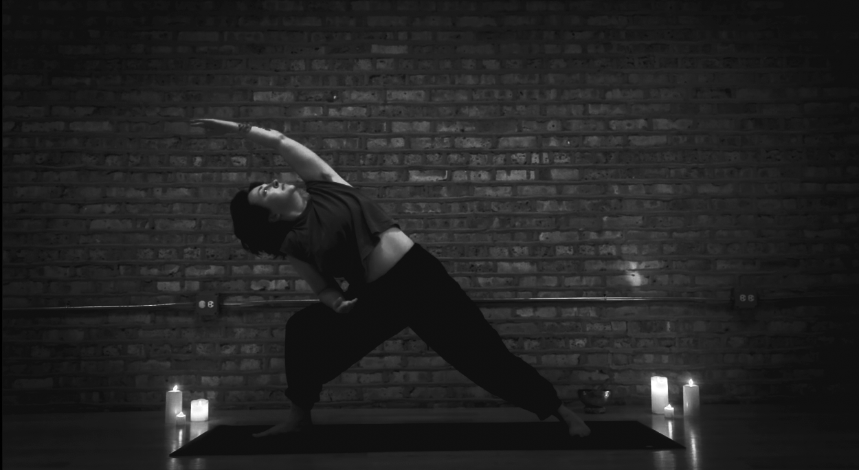A woman practicing yoga in a dimly lit room with candles and a brick wall background.