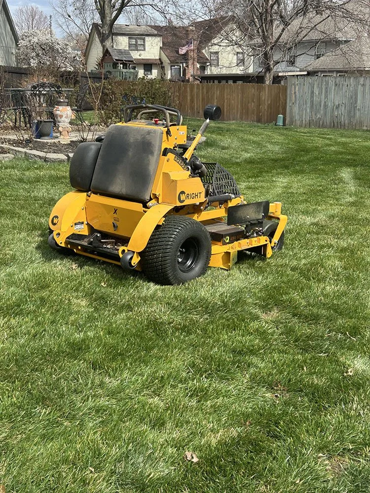 A large yellow Wright stand-on lawn mower on a green grassy yard, with a wooden fence, trees, and houses in the background.
