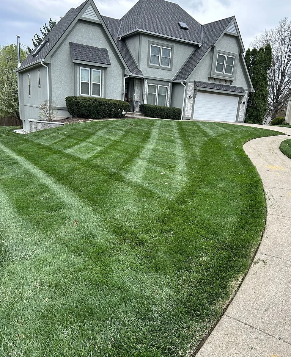 A two-story house with gray siding, a dark gray roof, and a front yard with freshly mowed grass showing checkered mowing patterns. There is a curved sidewalk leading to the front entrance and a white garage door.