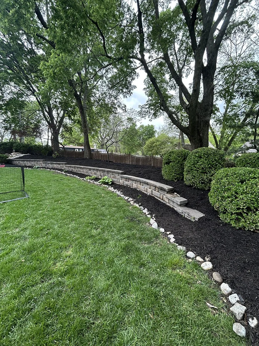 A landscaped backyard with a green lawn, a stone retaining wall, and neatly trimmed bushes under large trees with green foliage.