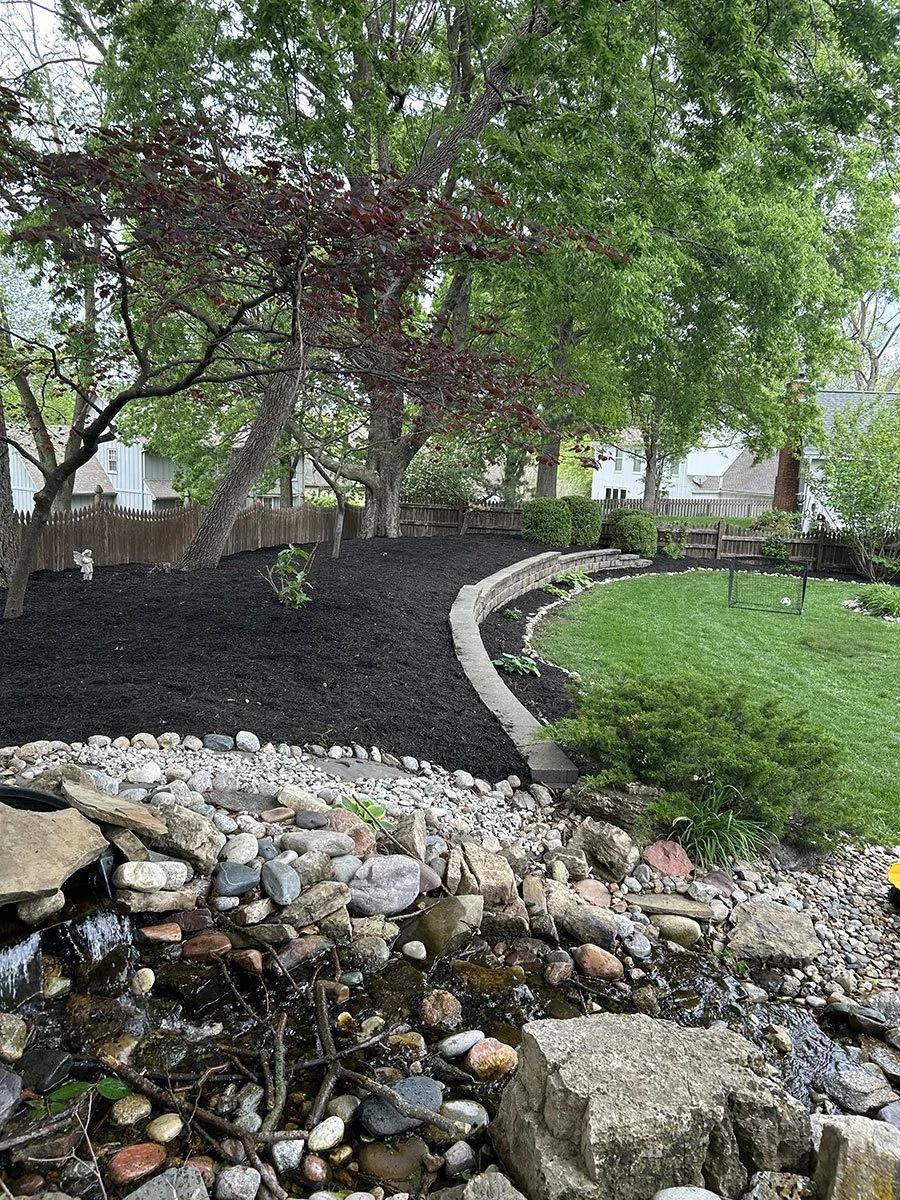 A landscaped backyard with a small pond filled with rocks and water in the foreground, green grass, bushes, and trees, with a curved stone retaining wall and a wooden fence in the background.