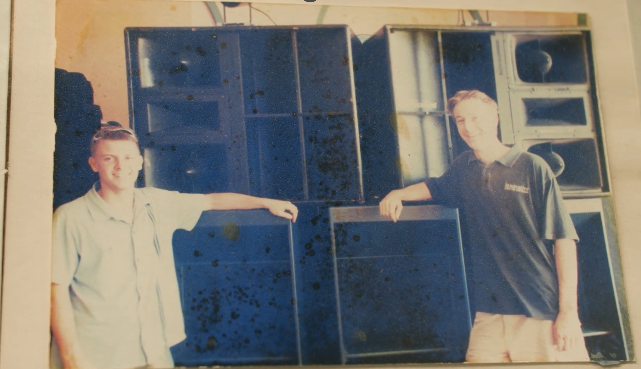 Two young men smiling and posing in front of large black speakers, one wearing a light-colored shirt and the other wearing a dark polo shirt with logos.