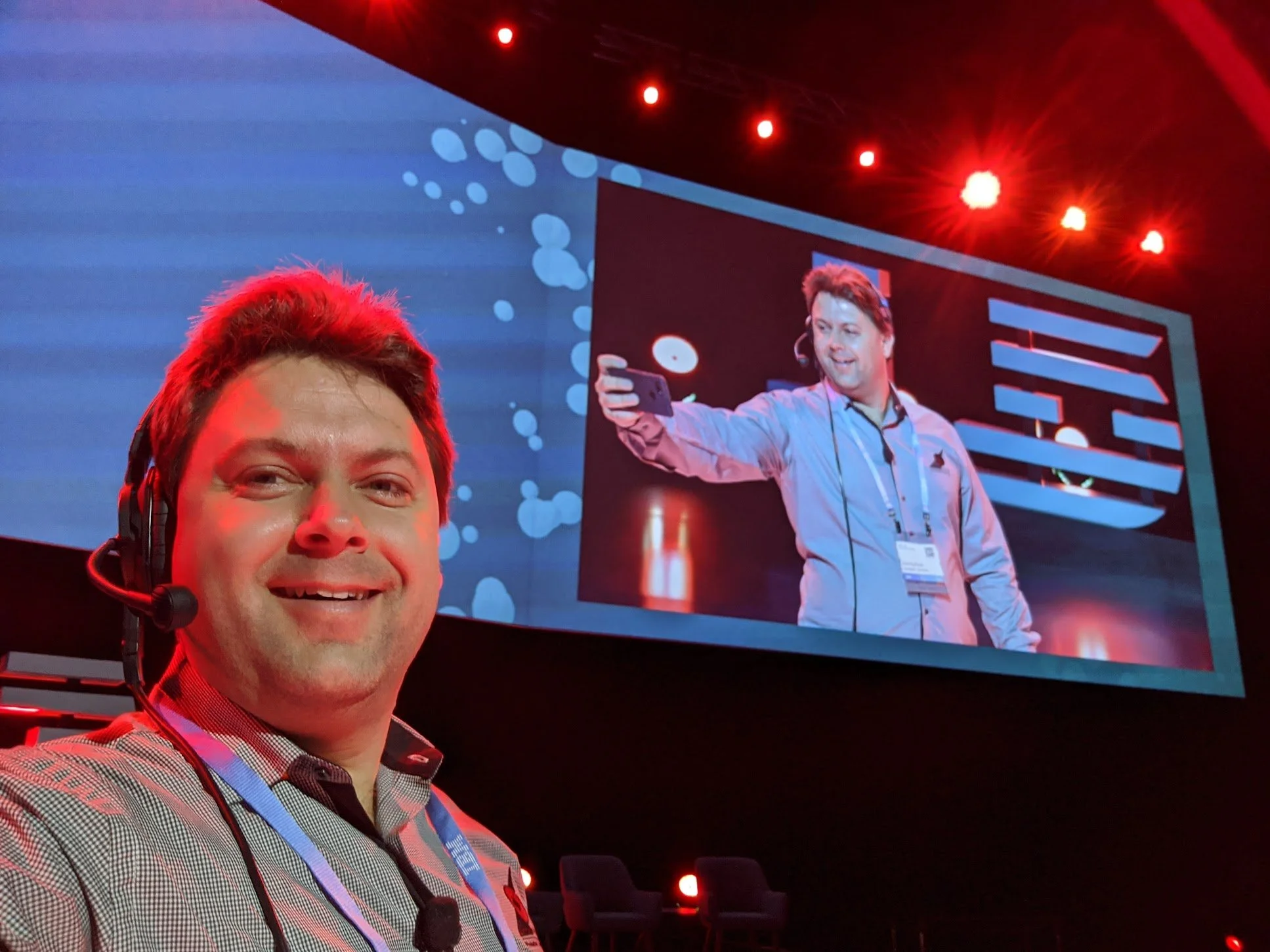 A man with a headset taking a selfie at a conference with a large screen behind him showing another man on stage speaking or presenting.