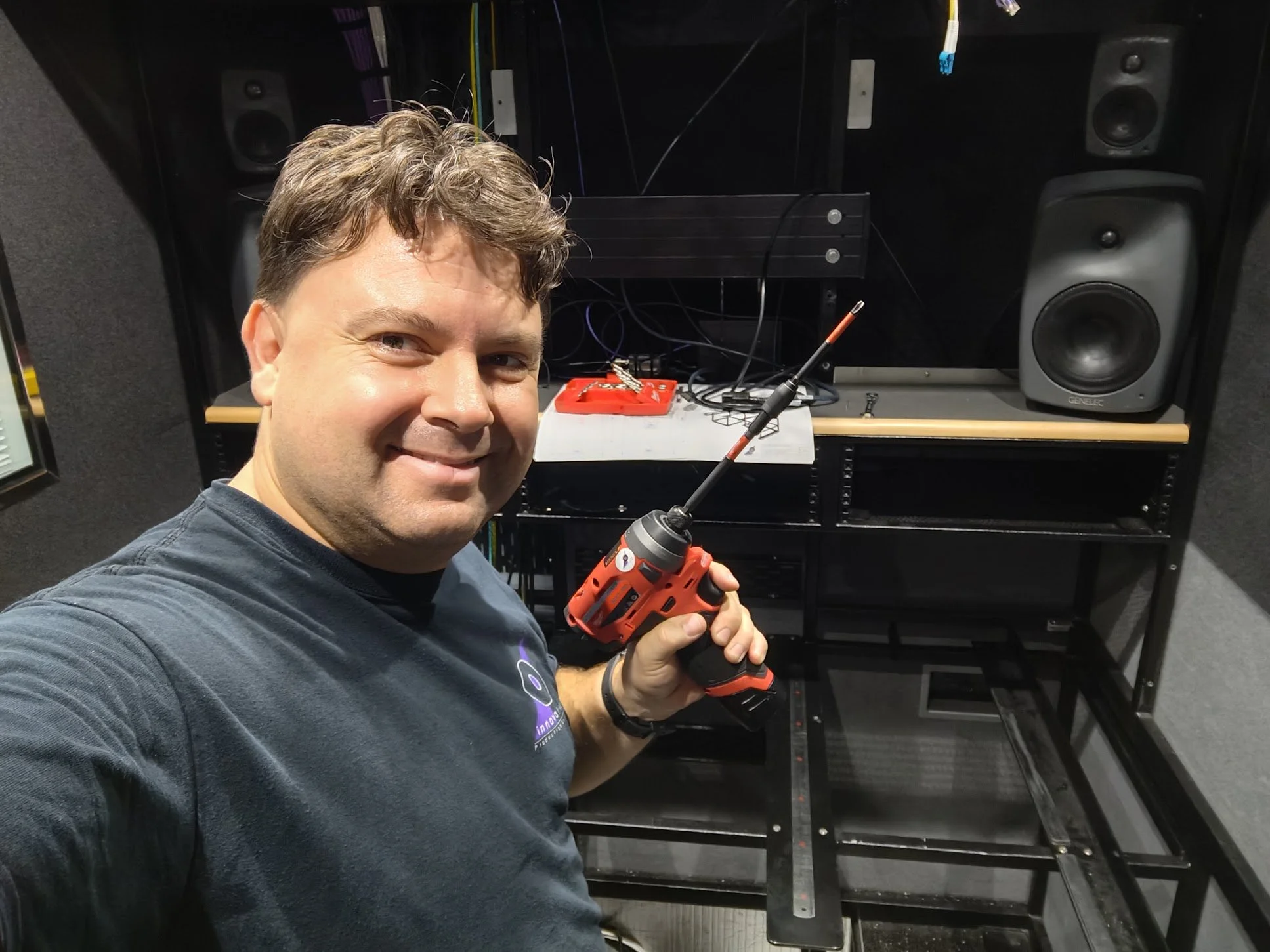 A smiling man holding a cordless drill in a workspace with sound equipment, including two large speakers and various tools and wires on a black shelving unit.