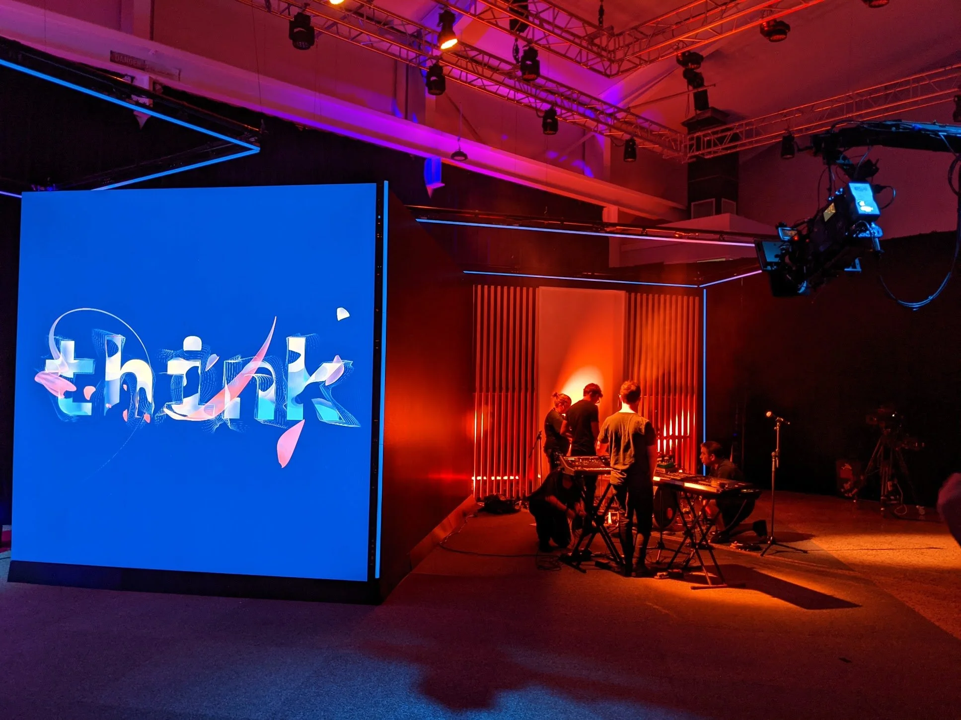 A stage setup with a large blue screen displaying the word 'think' and a stylized swoosh. There are musicians preparing with electronic instruments under orange and purple stage lighting. A camera crane hangs from the ceiling.