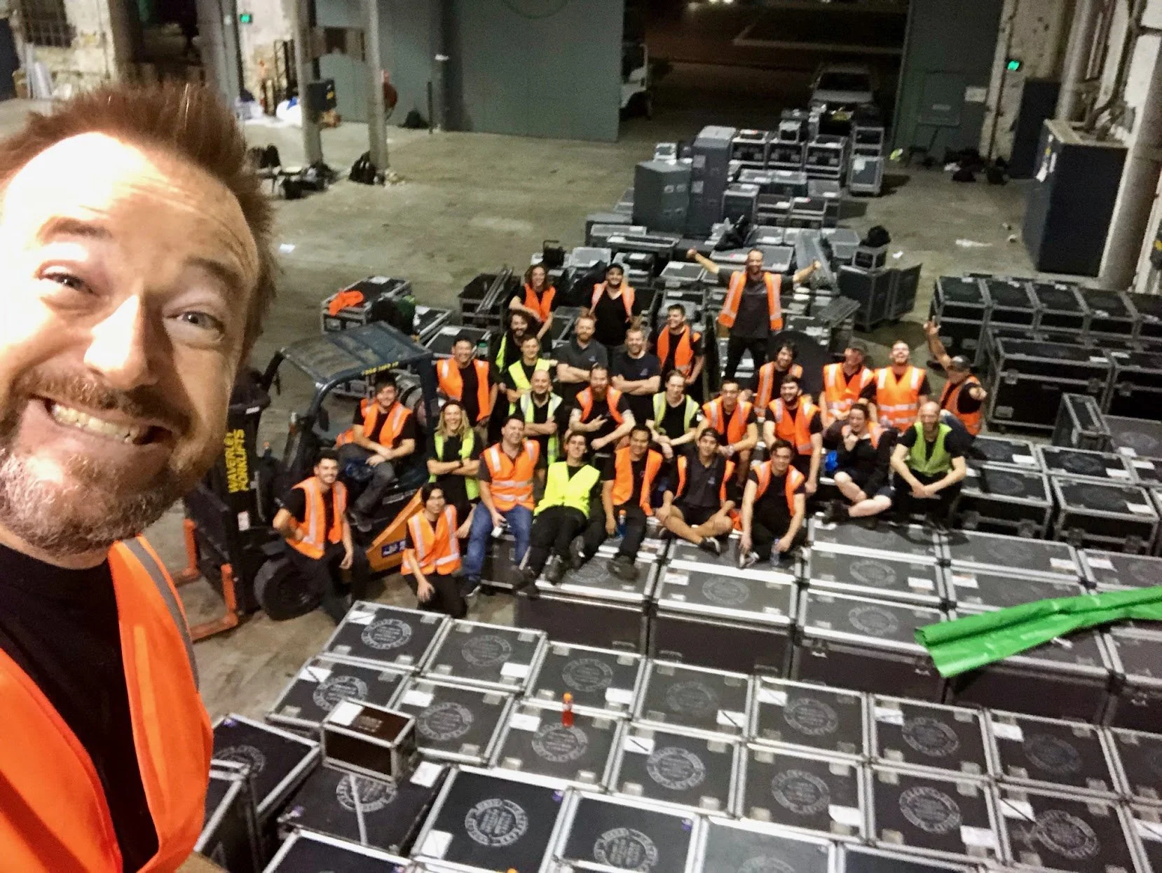 A man with a beard and orange safety vest taking a selfie with a group of workers in orange and yellow safety vests in a warehouse or storage area filled with black cases and equipment.