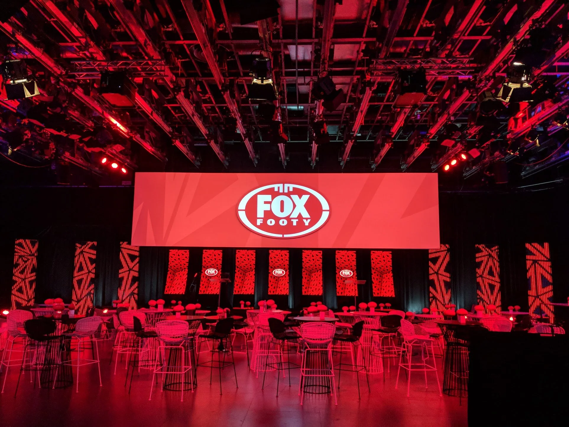 Event space with tables, chairs, and large screen displaying 'FOX FOOTY' logo, illuminated with red and black lighting.
