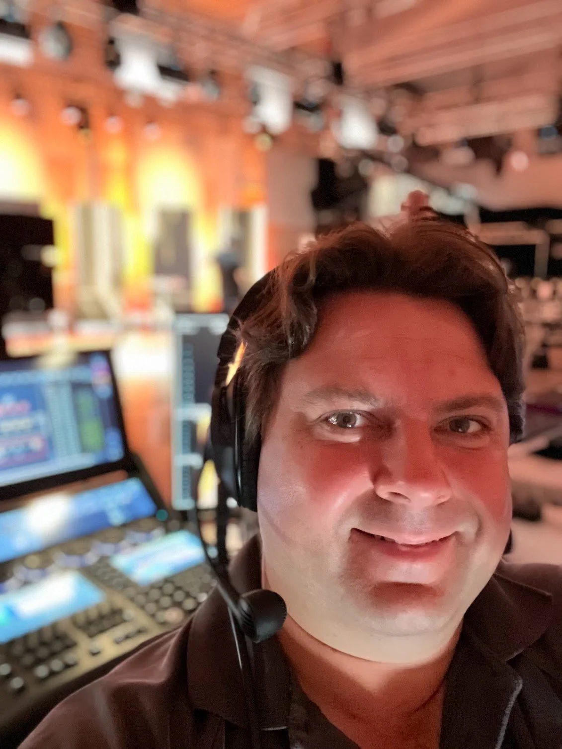 A person with dark hair wearing a headset, taking a selfie in a room with multiple screens and audio equipment in the background.