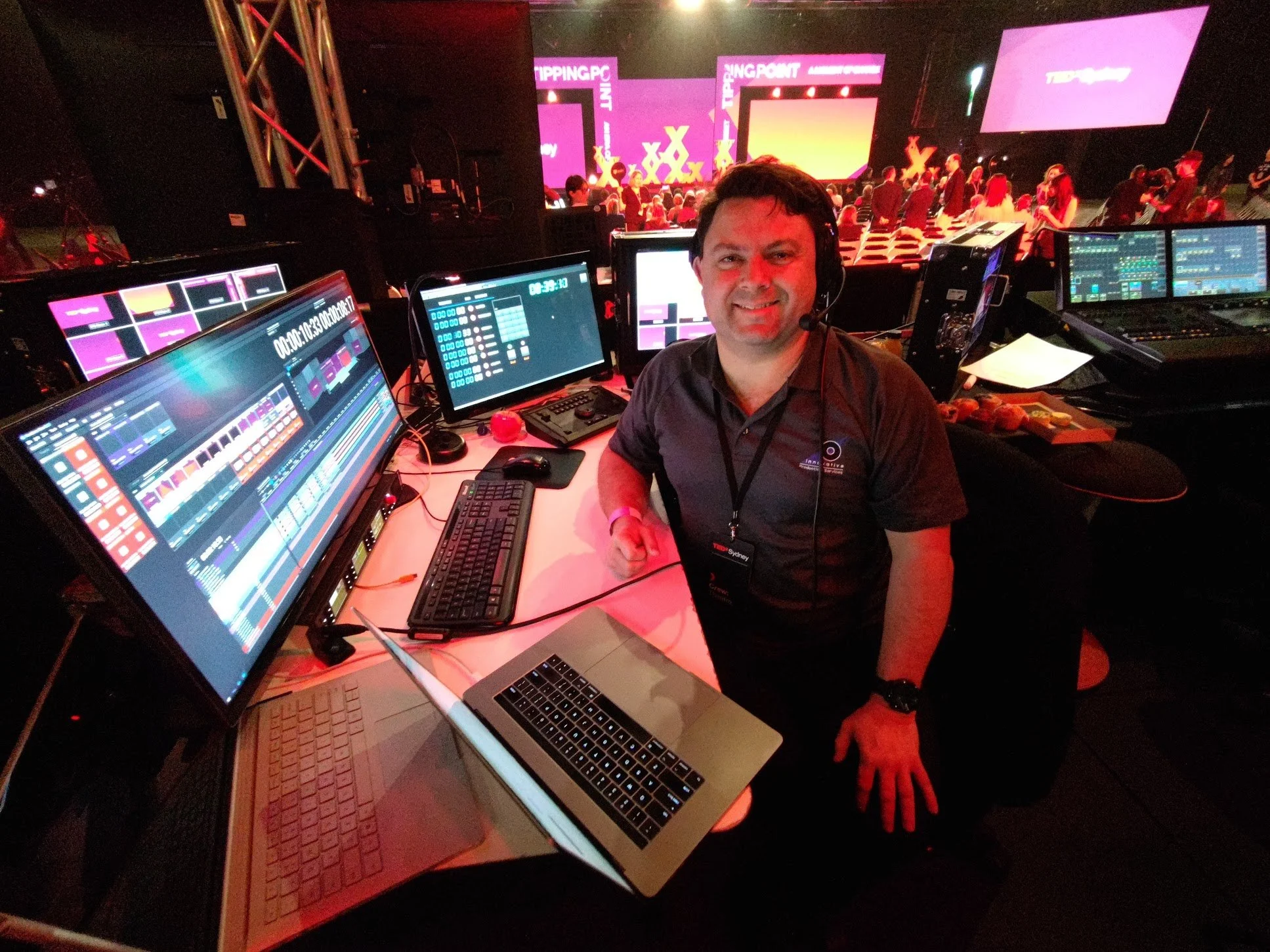 A man in a black shirt sitting at a control desk with multiple monitors displaying audio and video editing software, smiling at the camera, with a stage and audience visible in the background.