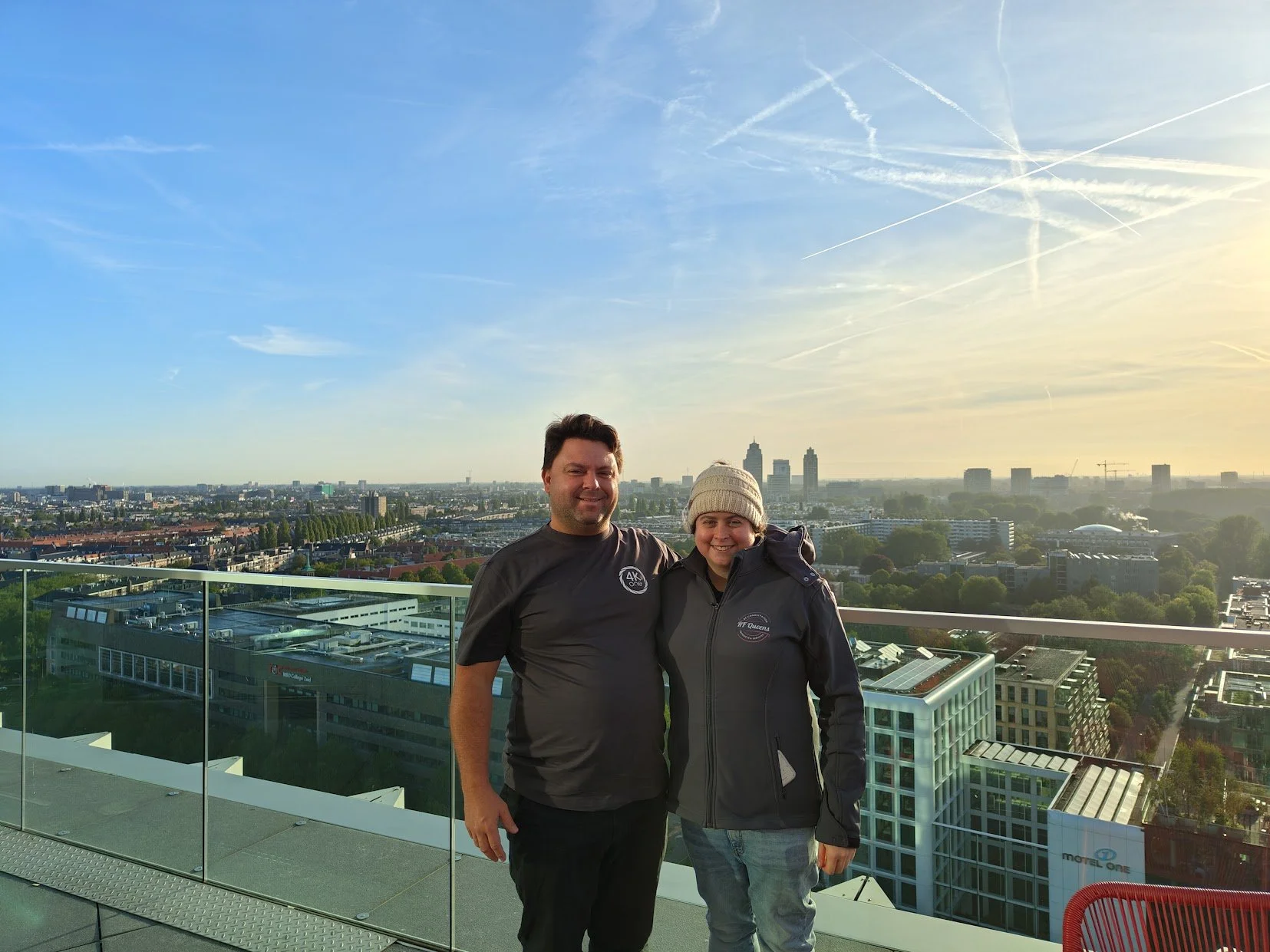 A man and woman standing together on a rooftop with a city skyline in the background during sunset, both smiling.