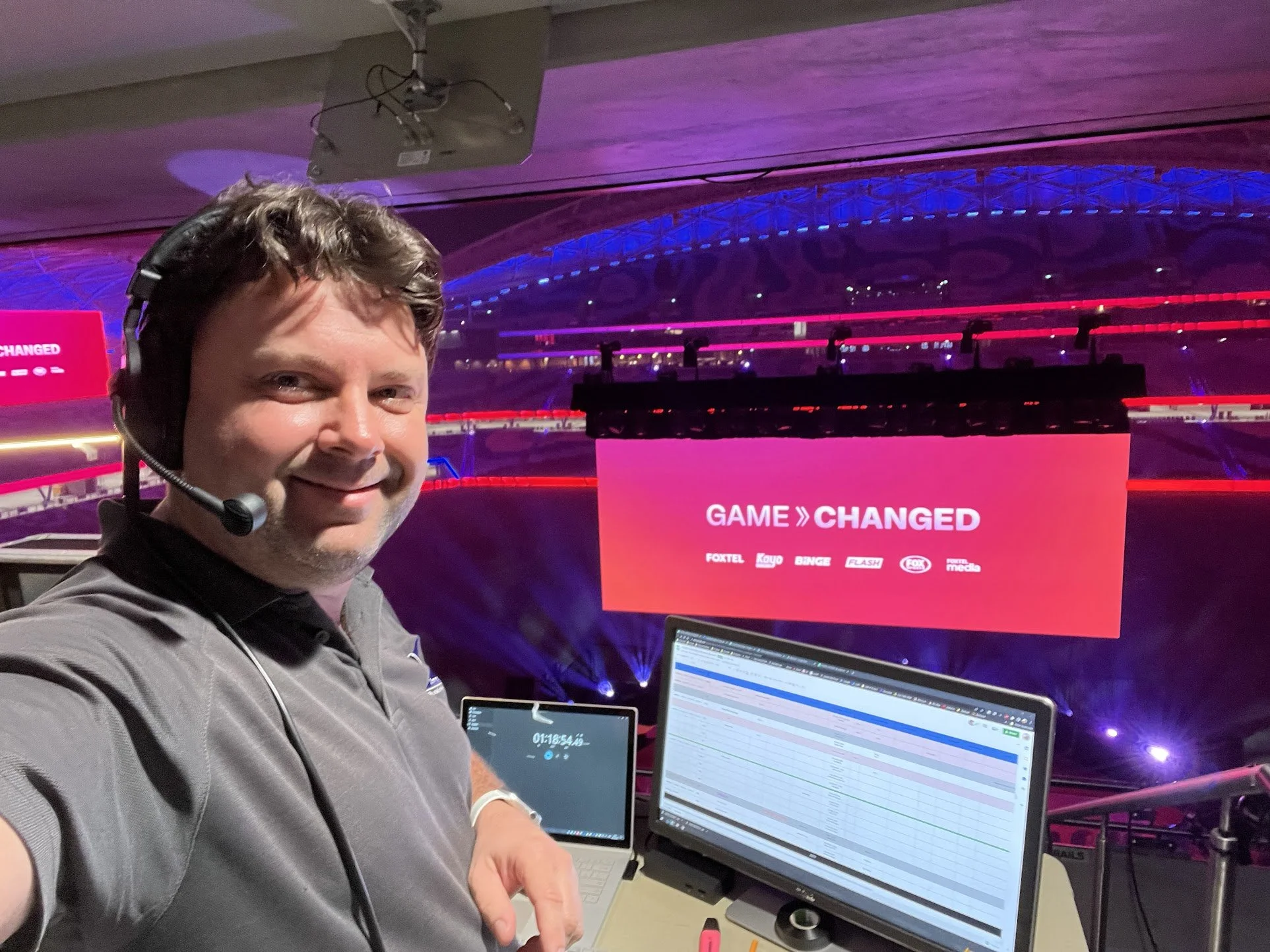 A man wearing a headset takes a selfie in an arena with a stage and large screen displaying 'GAME CHANGED' in the background. He is seated near a laptop and an iPad, both showing schedules or data.
