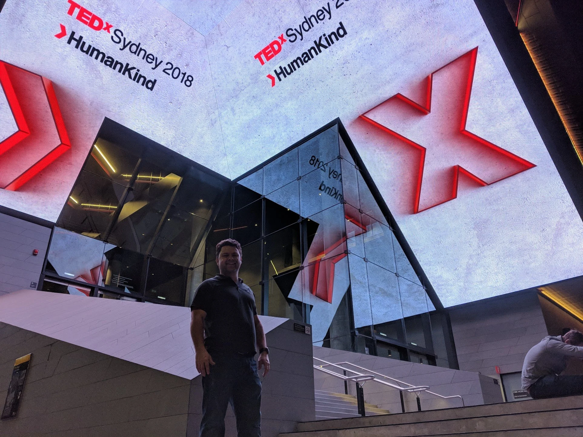A man standing in front of a large digital screen at TEDx Sydney 2018 event, with the TEDx logo and event details displayed.