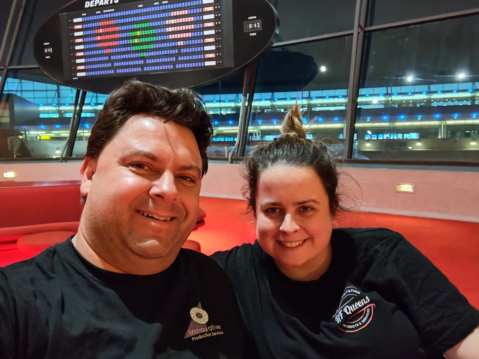 A man and woman smiling and taking a selfie at an airport. The man is on the left, wearing a black shirt with a logo that reads 'Innovative Production Services.' The woman is on the right, wearing a black shirt with a logo that reads 'RF Queens.' Beh