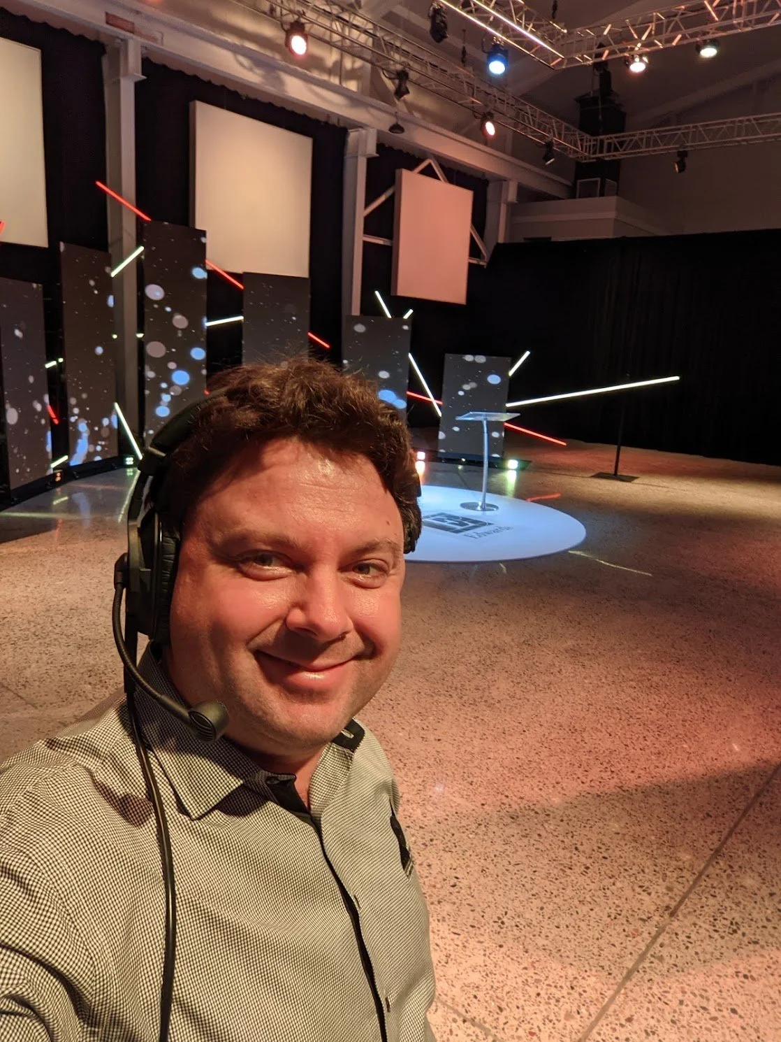 Smiling man with a headset taking a selfie in an auditorium or conference stage setup with geometric light decorations and a podium in the background.