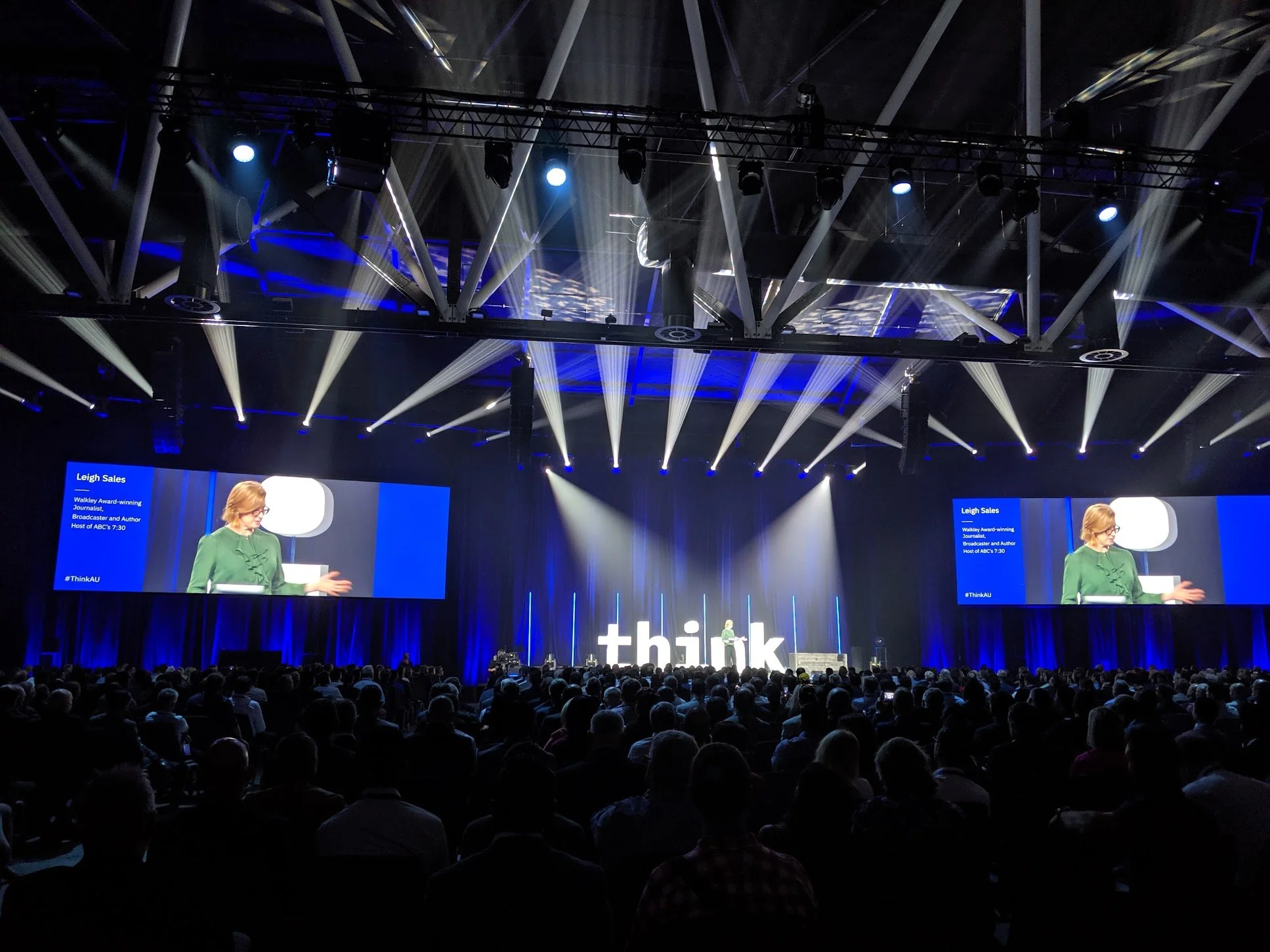 Large audience attending a conference with a stage and two large screens displaying a woman speaking. The stage has bright lighting and a 'think' sign at the back.