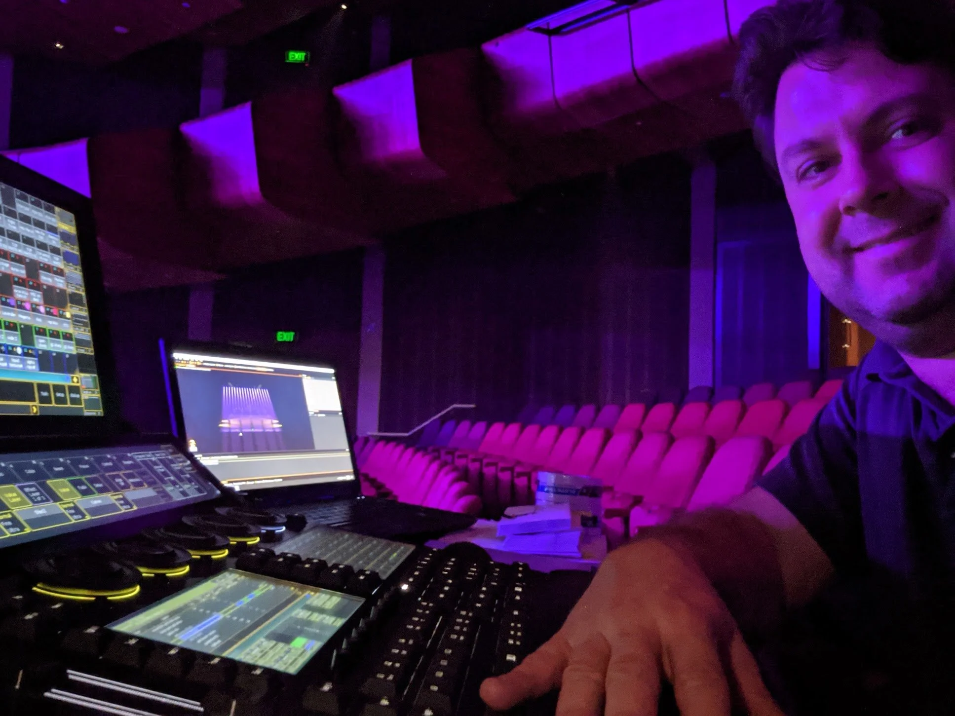 A man smiling at a sound and lighting control booth with multiple monitors and equipment in a dark, purple-lit theater or auditorium.