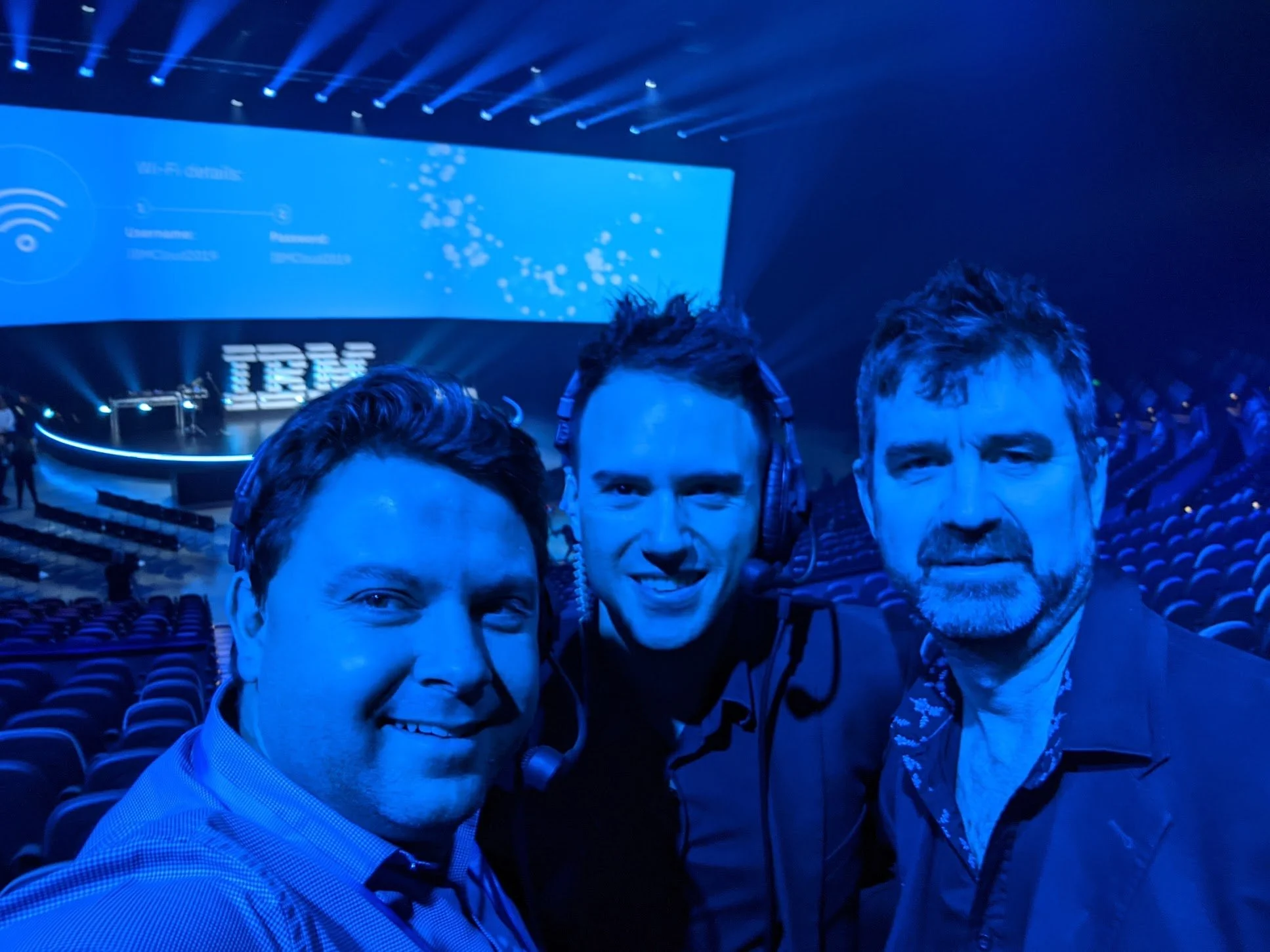 Three men taking a selfie at a tech conference or event, with a large screen and IBM logo in the background.