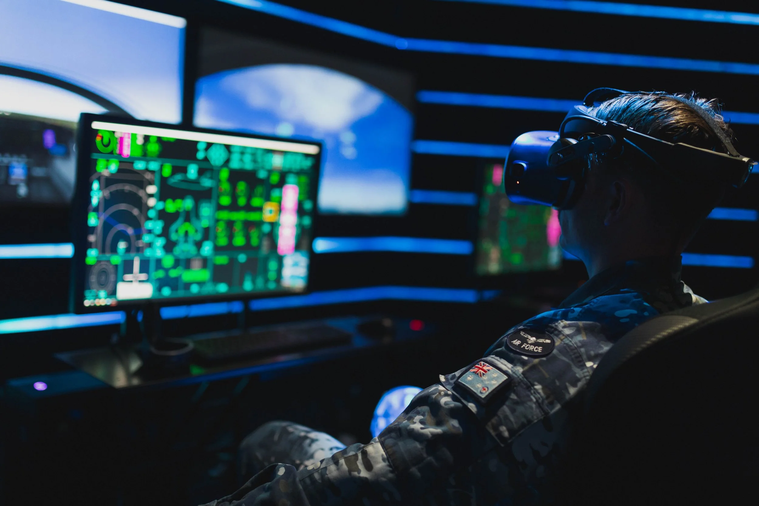 Person wearing military camouflage uniform with British flag patch and 'Air Force' badge, sitting in front of multiple computer screens displaying digital graphics and radar interfaces, in dimly lit room with blue lighting.