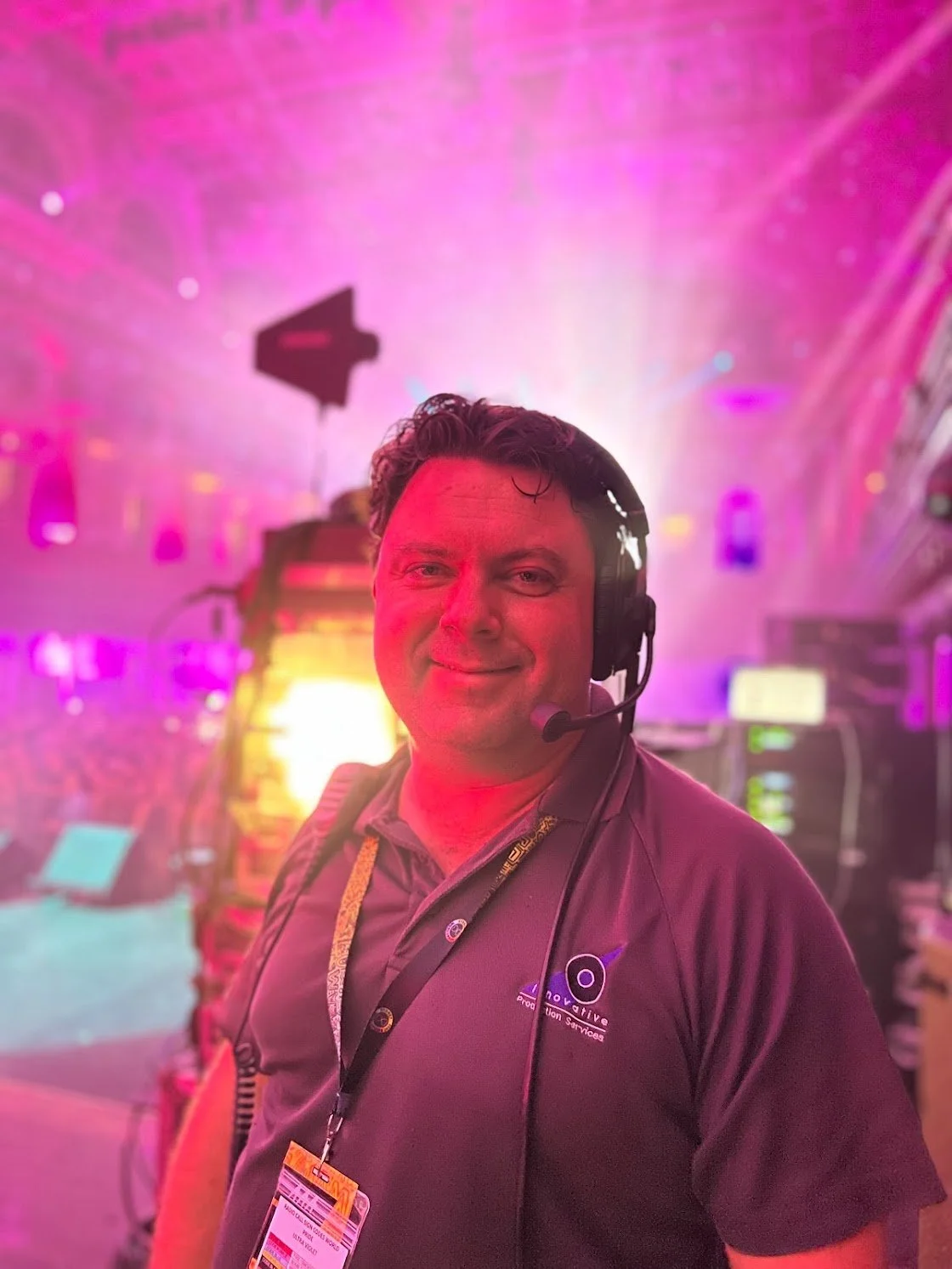 A man wearing a headset and a gray polo shirt with a lanyard around his neck, smiling at the camera against a vibrant, colorful background with stage equipment and pink lighting.