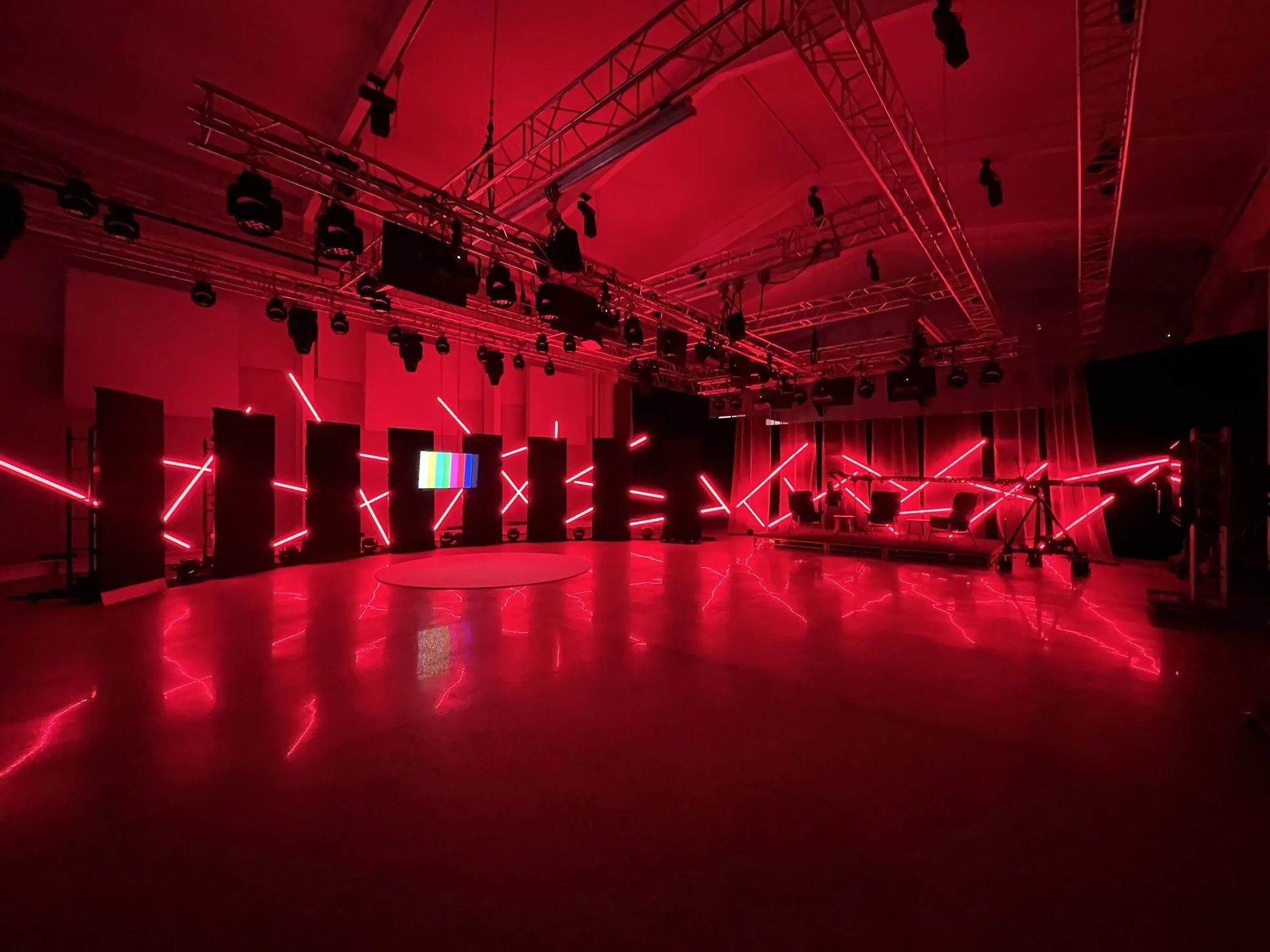 An indoor stage setup with a red lighting theme, featuring multiple vertical black panels, red laser or LED light beams crossing in various directions, and a flat circular platform on the floor. There is a large screen with color bars at the center b
