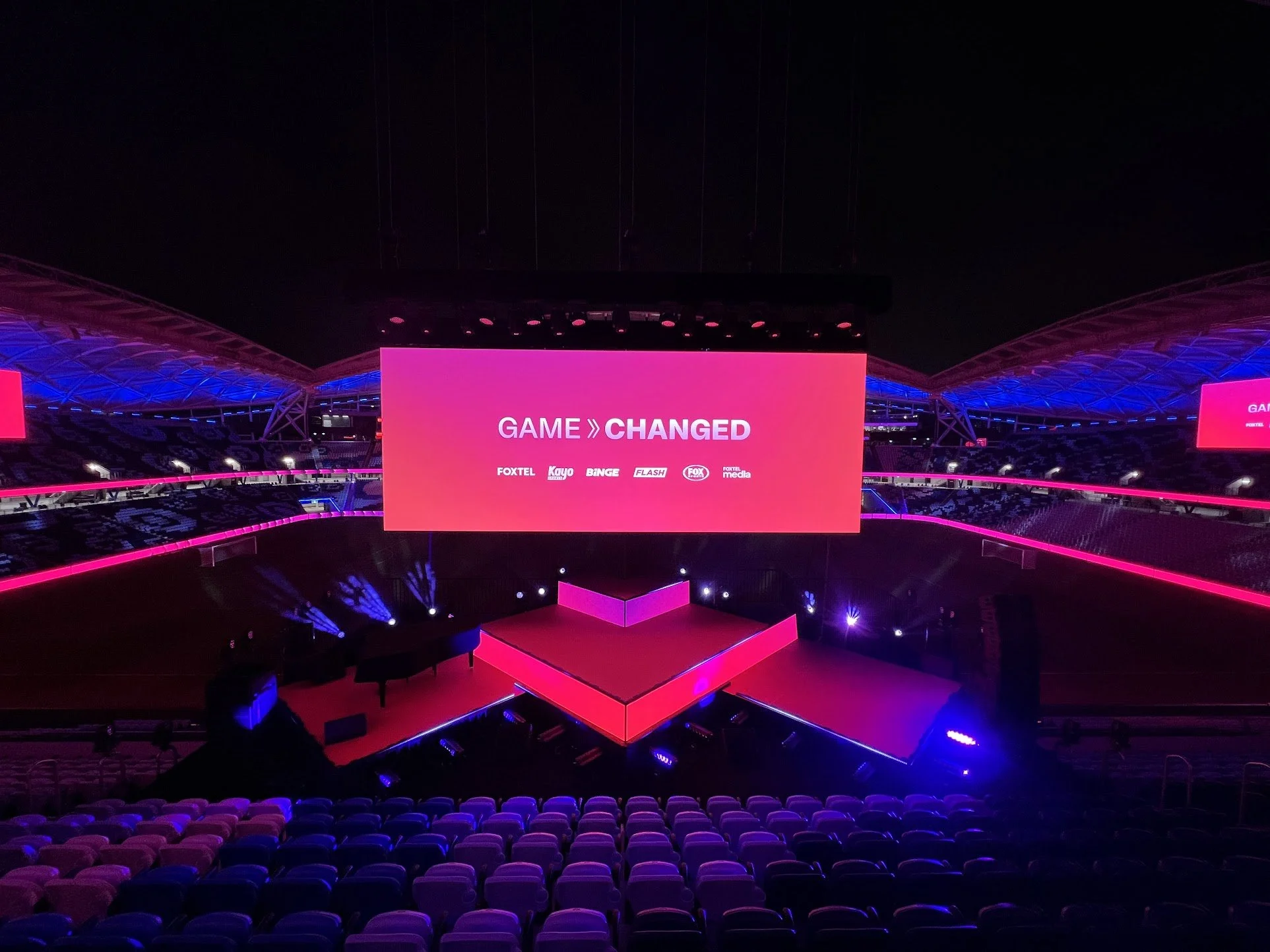 Empty indoor stadium with large pink LED screen reading 'Game Changed' and logos of media companies, illuminated with pink and purple lighting.