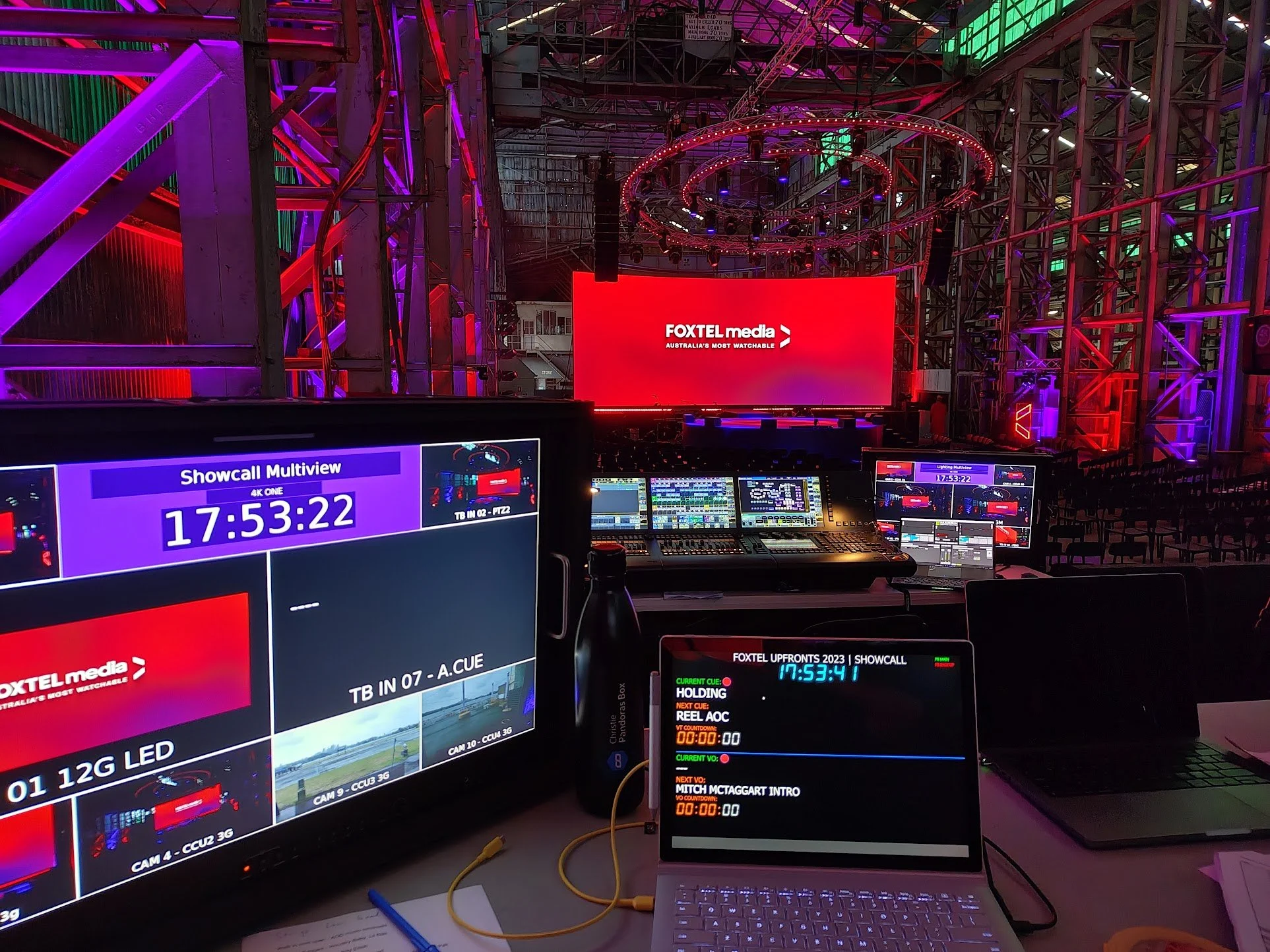 A control room with multiple screens displaying live feeds, timers, and technical information at an event for Foxtel media, with lighting equipment and industrial steel framework in the background.