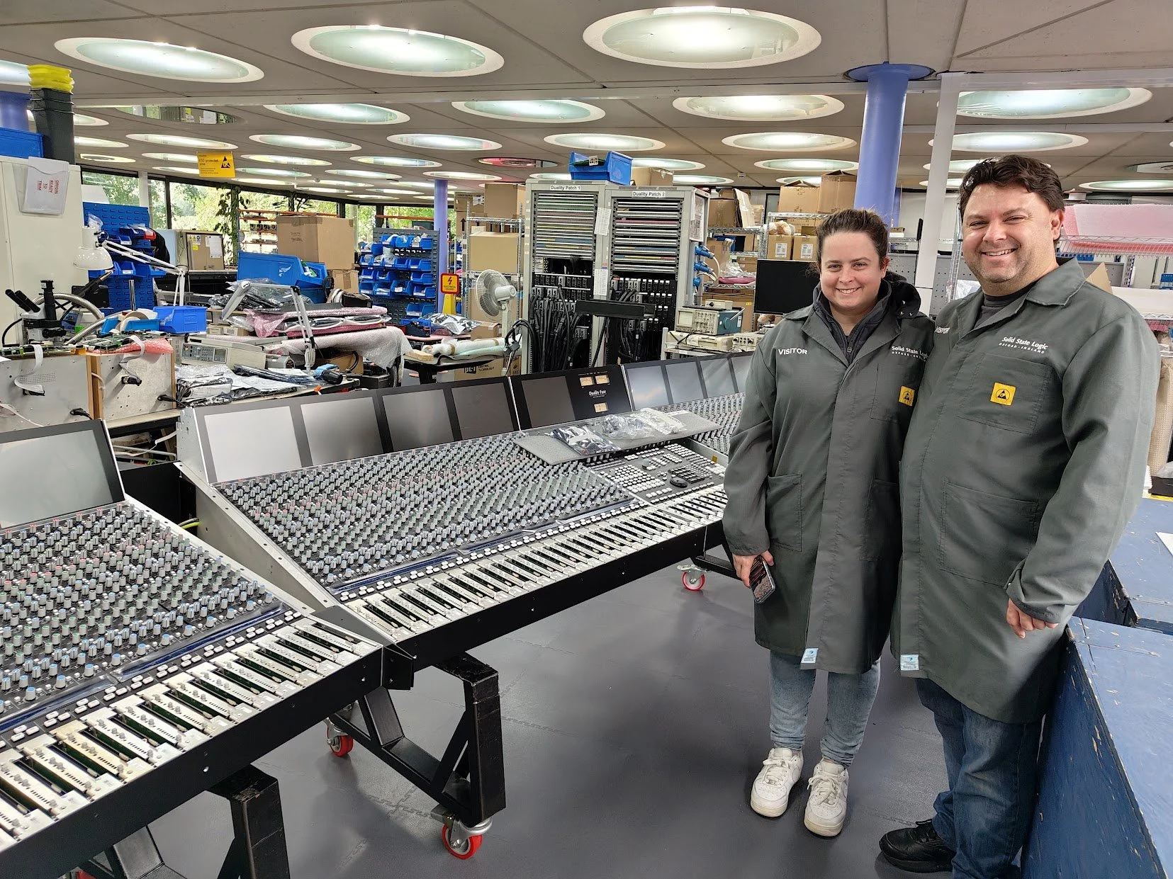 Two technicians in gray uniforms standing next to a large audio mixing console in an industrial workspace with tools, equipment, and boxes in the background.