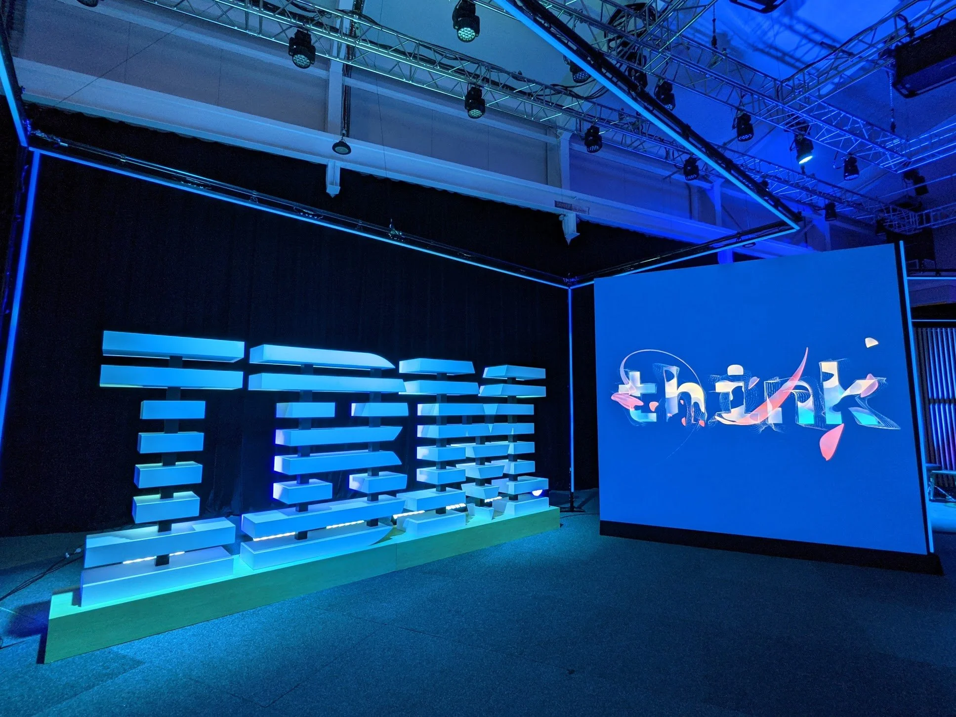 Stage setup with a large illuminated text sign and a digital screen displaying the word 'think' with colorful abstract graphic elements, in a conference or event space.